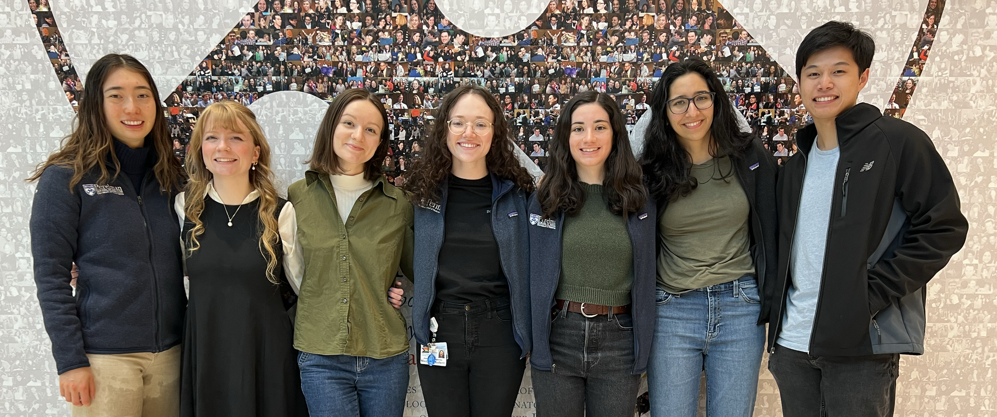 A group photo of medical students Catherine Shen, Elizabeth Whidden, Phoebe Cunningham, Jess Campanile, Sarah Wornow, Isha Thapar, and Jonathan Szeto standing in front of a photo collage in the shape of the Penn shield.