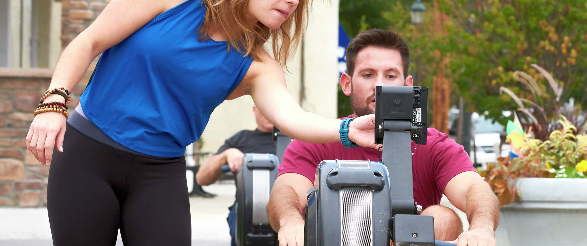 A female fitness instructor points at the controls of a rowing machine that a young man is using