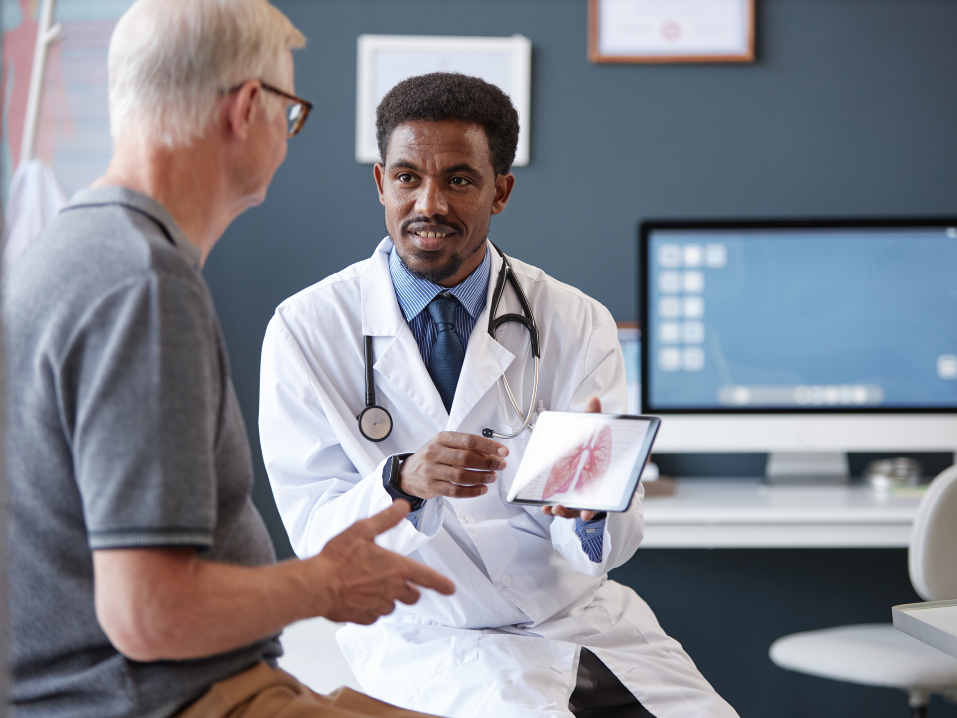  Doctor wearing lab coat and holding tablet while explaining treatment to senior patient during consultation in clinic.