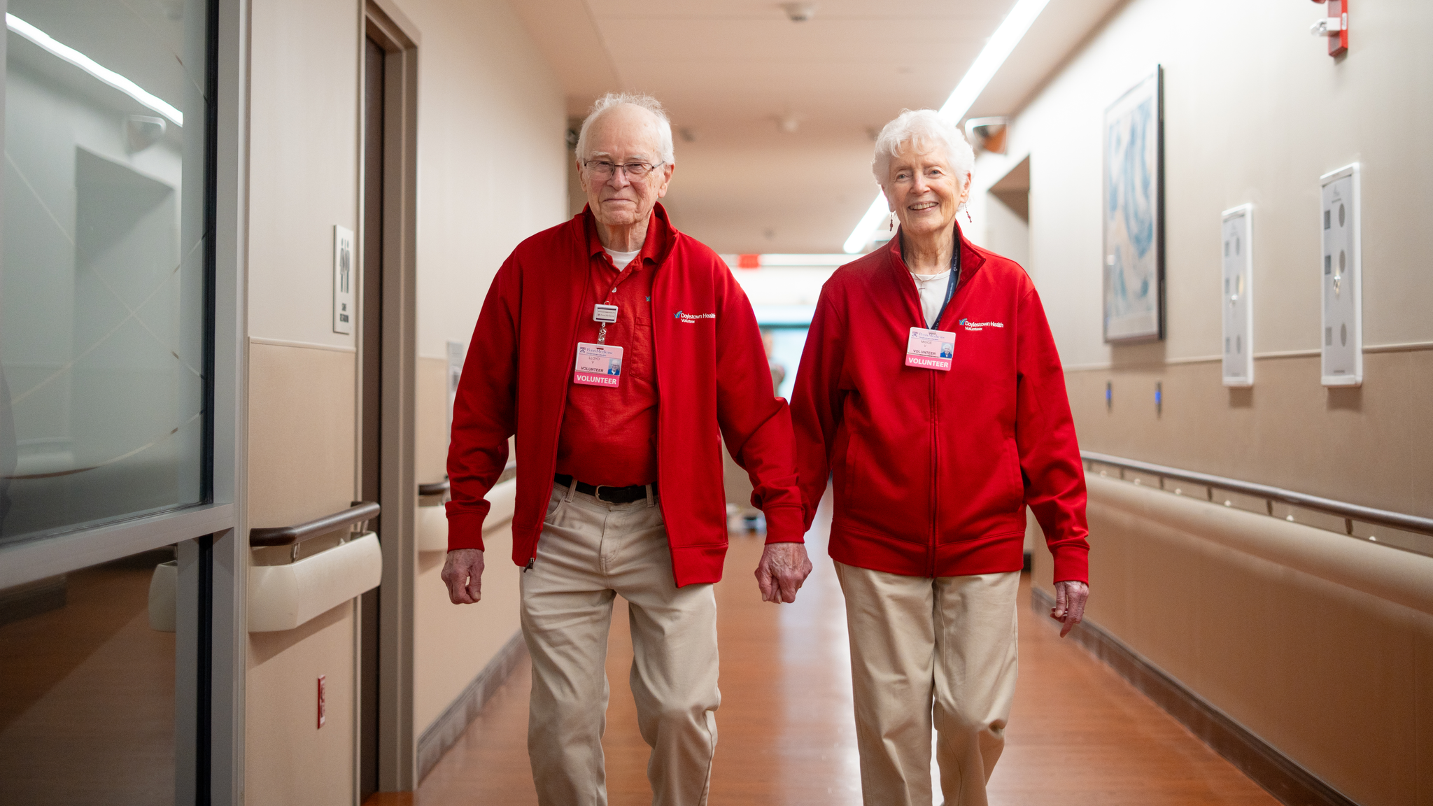 Lloyd and Midge smile at the camera as they walk hand-in-hand through a Doylestown Hospital hallway. They are wearing matching tan khaki pants and matching red sweatshirts