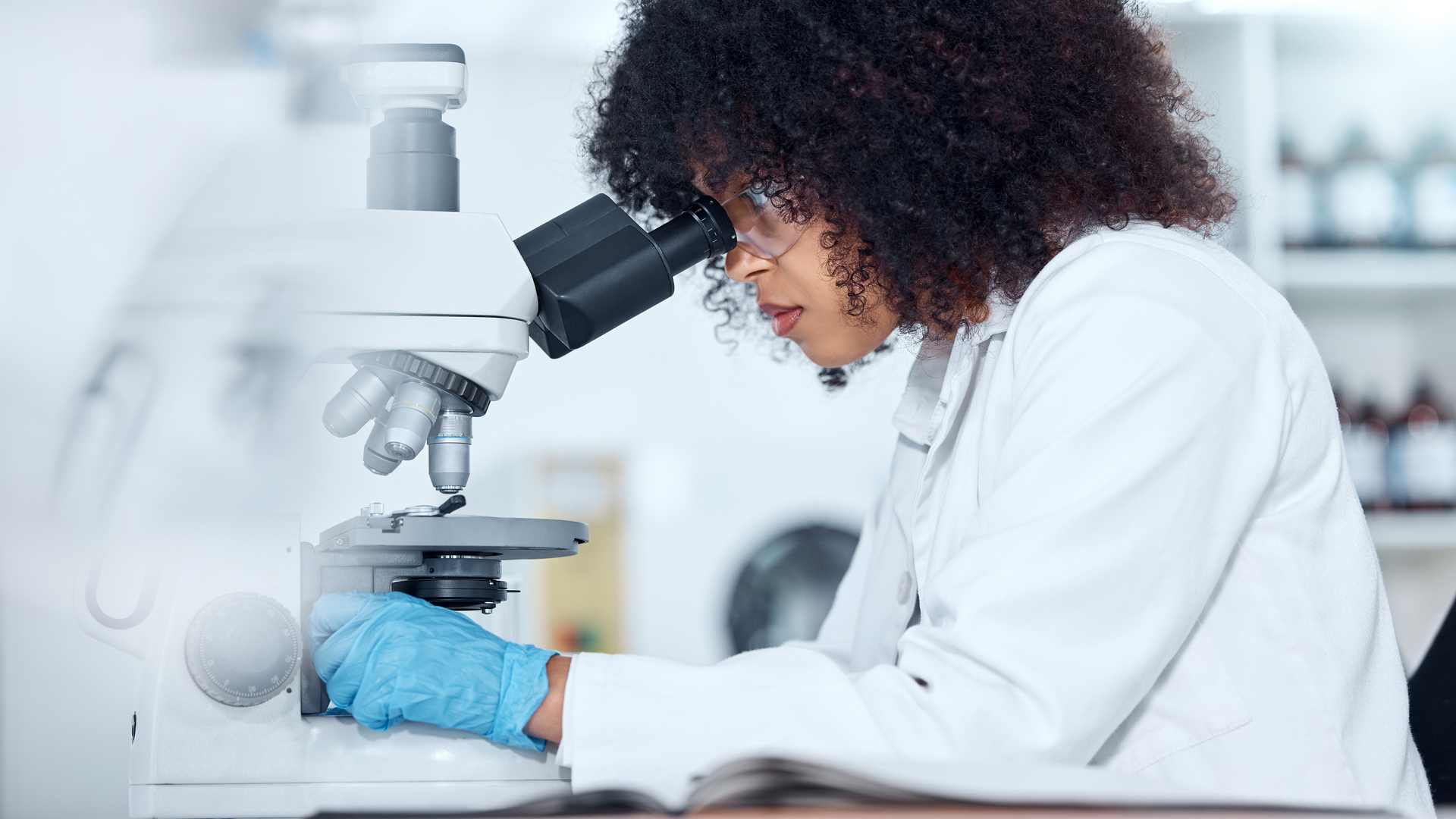 Scientist with curly hair wearing safety goggles and gloves analyzing medical test samples on a microscope in a lab.