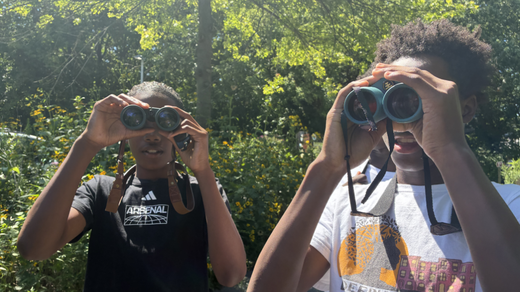 Two young people, one wearing a Deeply Rooted t-shirt, looking through binoculars in a wooded setting