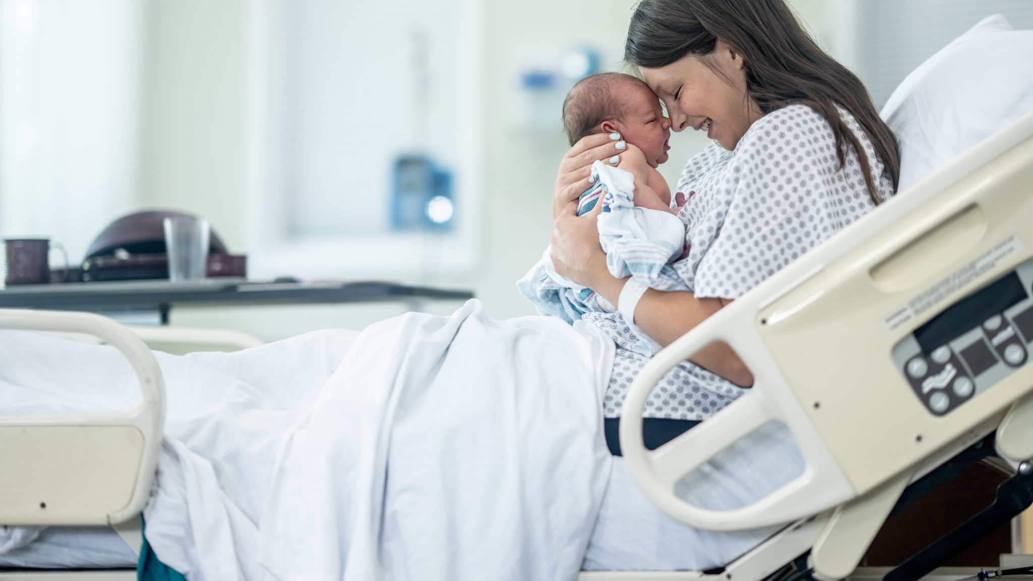 A new mother sits up in hospital bed shortly after delivery as she holds her newborn out in front of her 