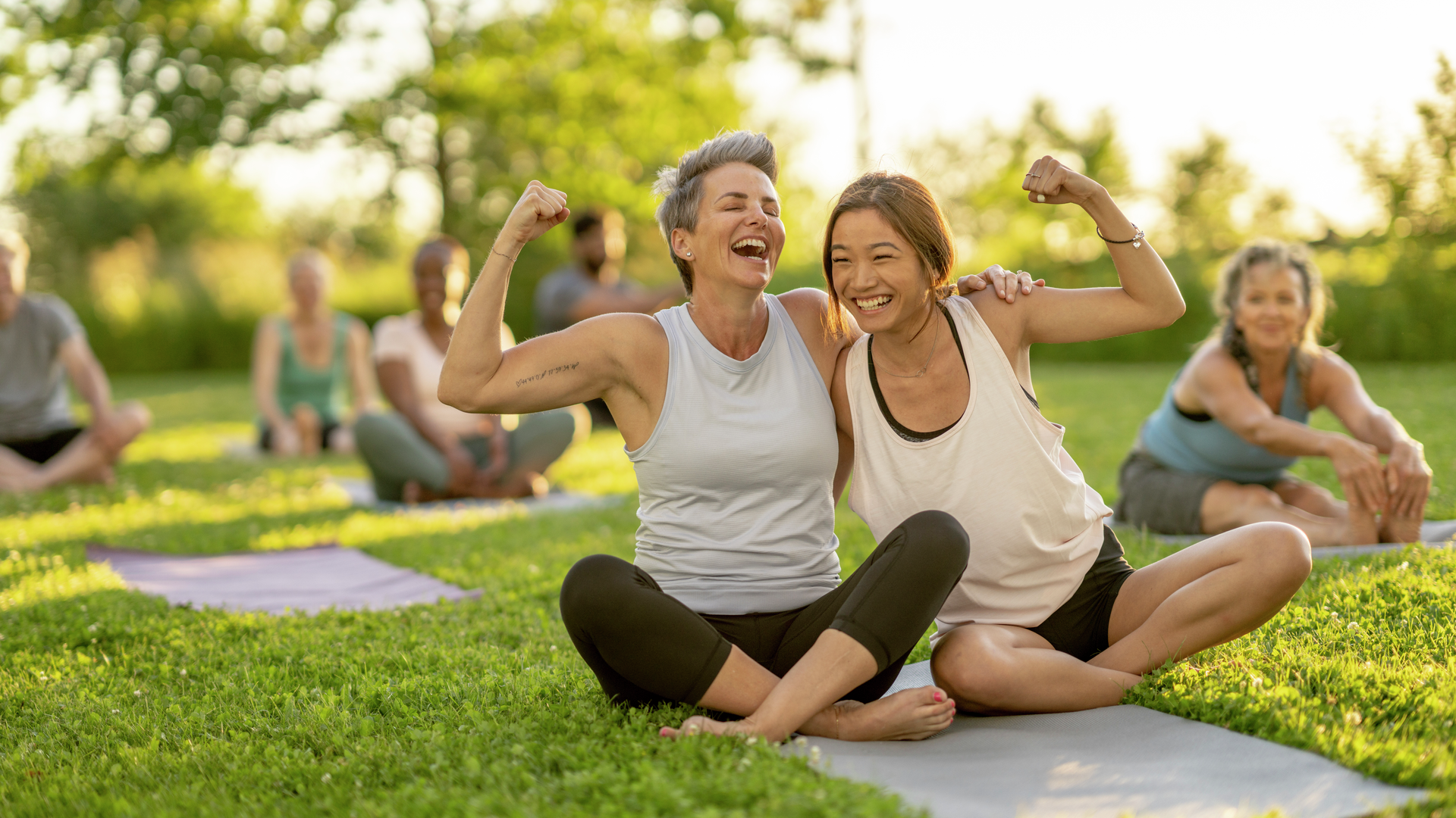 Two woman share a yoga mat as they lean in closely for a hug and flex their muscles