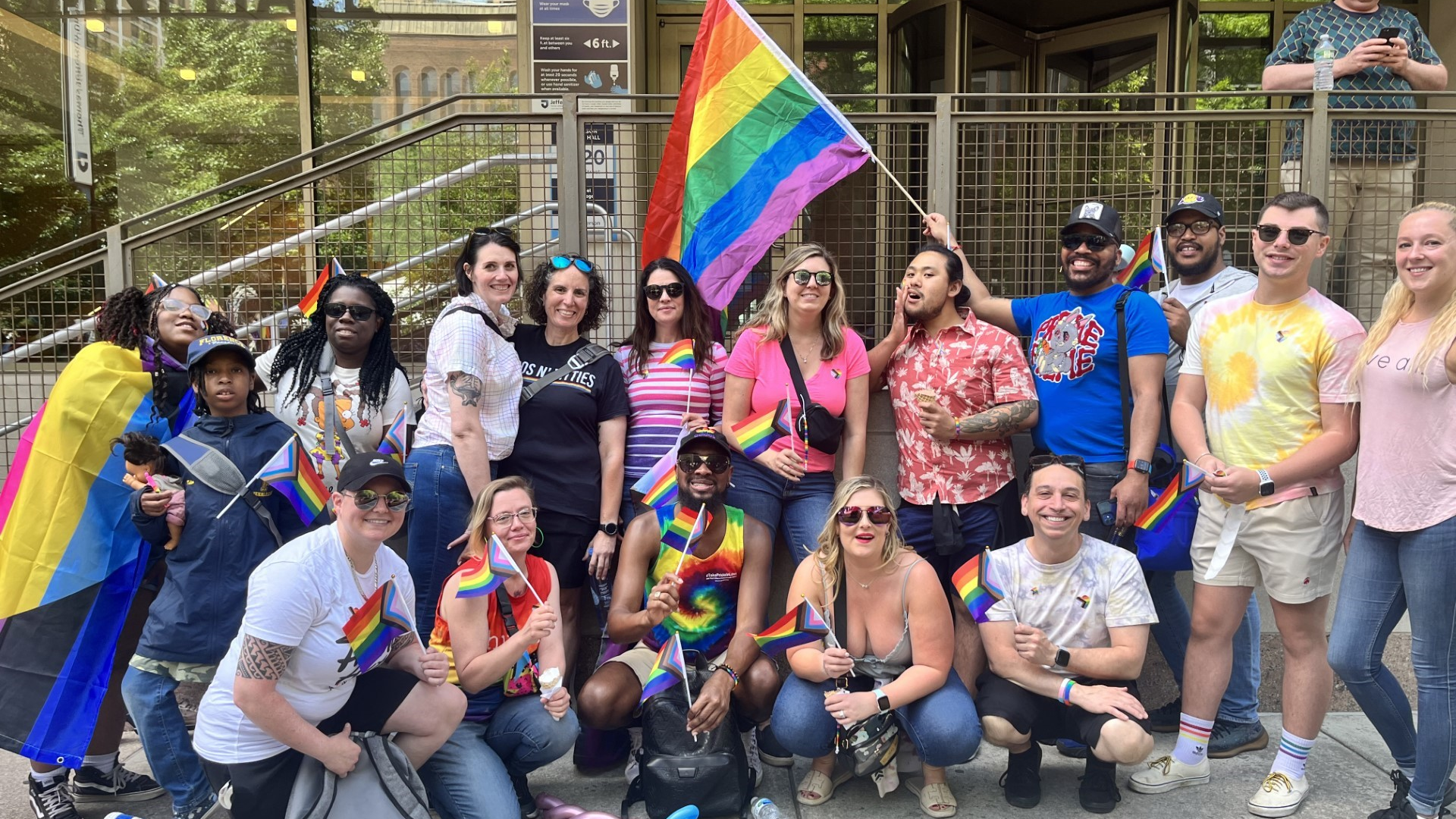 A group photo of about 20 people of diverse racial, ethnic, and gender presentations, most holding mini pride flags and one person waving a large flag