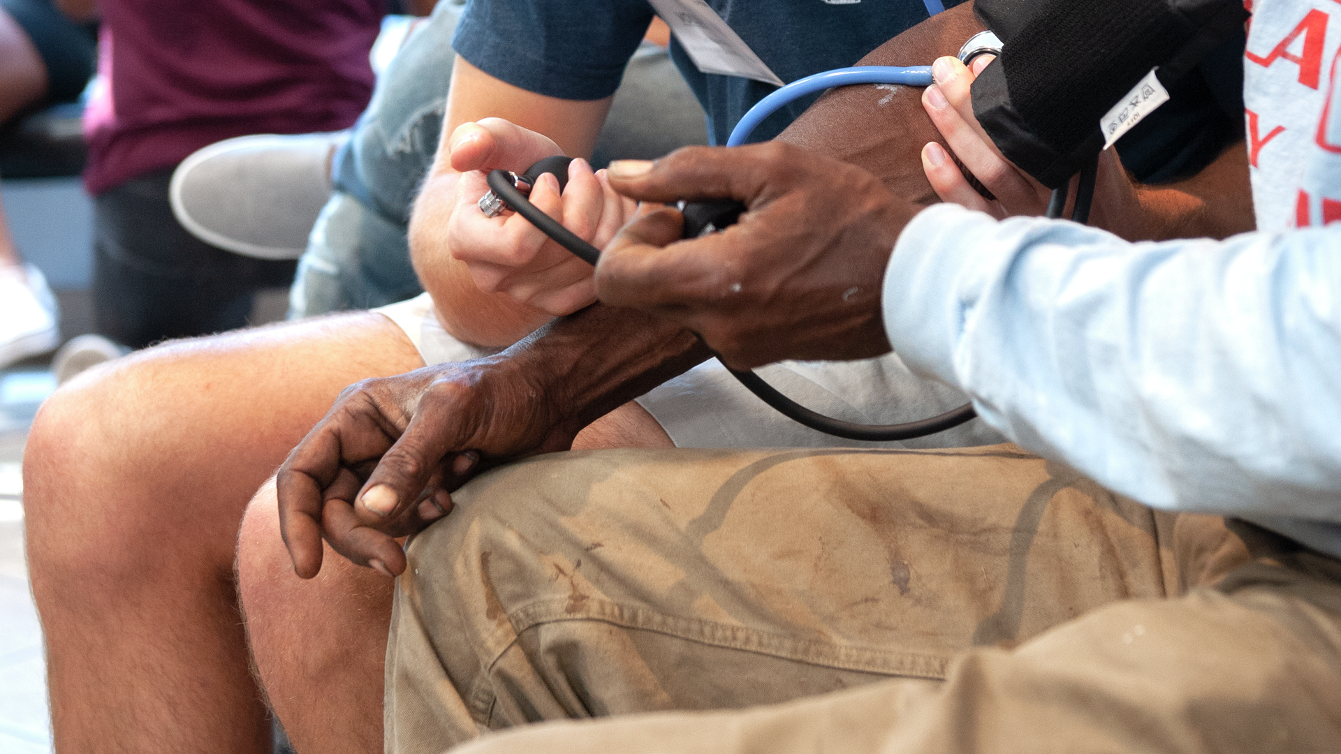 A medical student measures a Black man's blood pressure while other students and barbershop patrons can be seen in the background