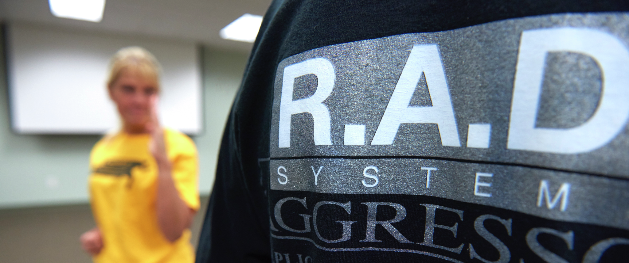 Back of a T-shirt saying "R.A.D. System" with out-of-focus Deborah Millar practicing self defense