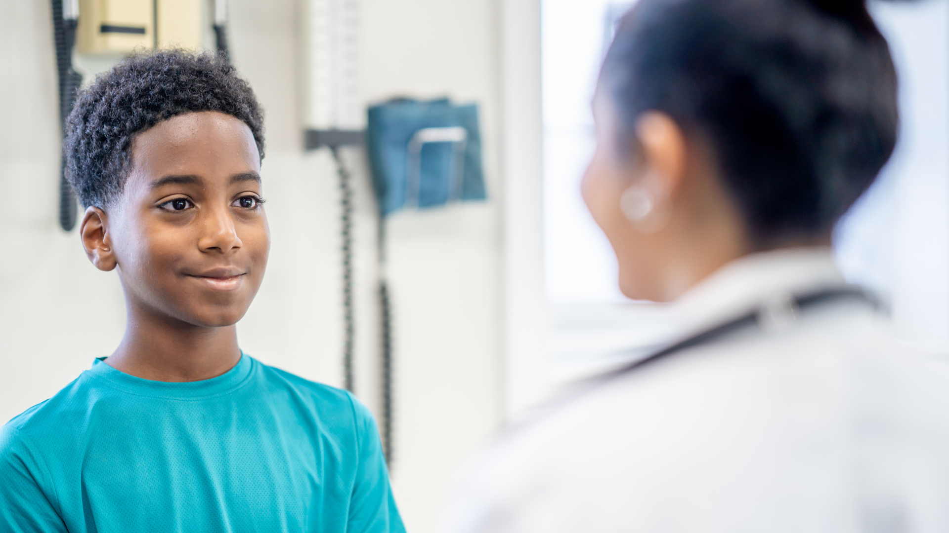 A young teen, of African decent, sits up on an exam table during a routine medical check-up. He is dressed casually and smiling as he talks with his doctor.