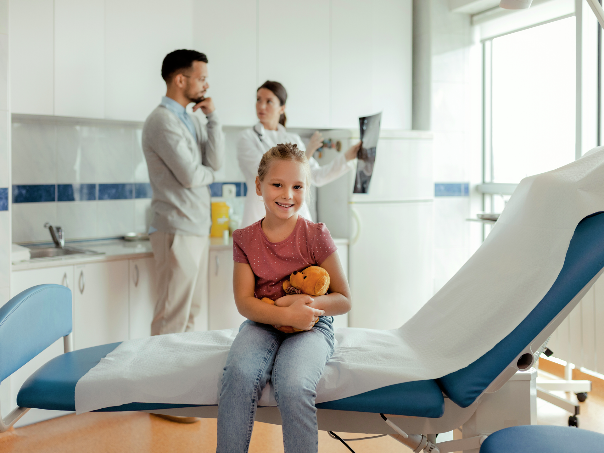 A young girl sitting on the examination table at a doctors appointment while her father and doctor stand behind her having a discussion.