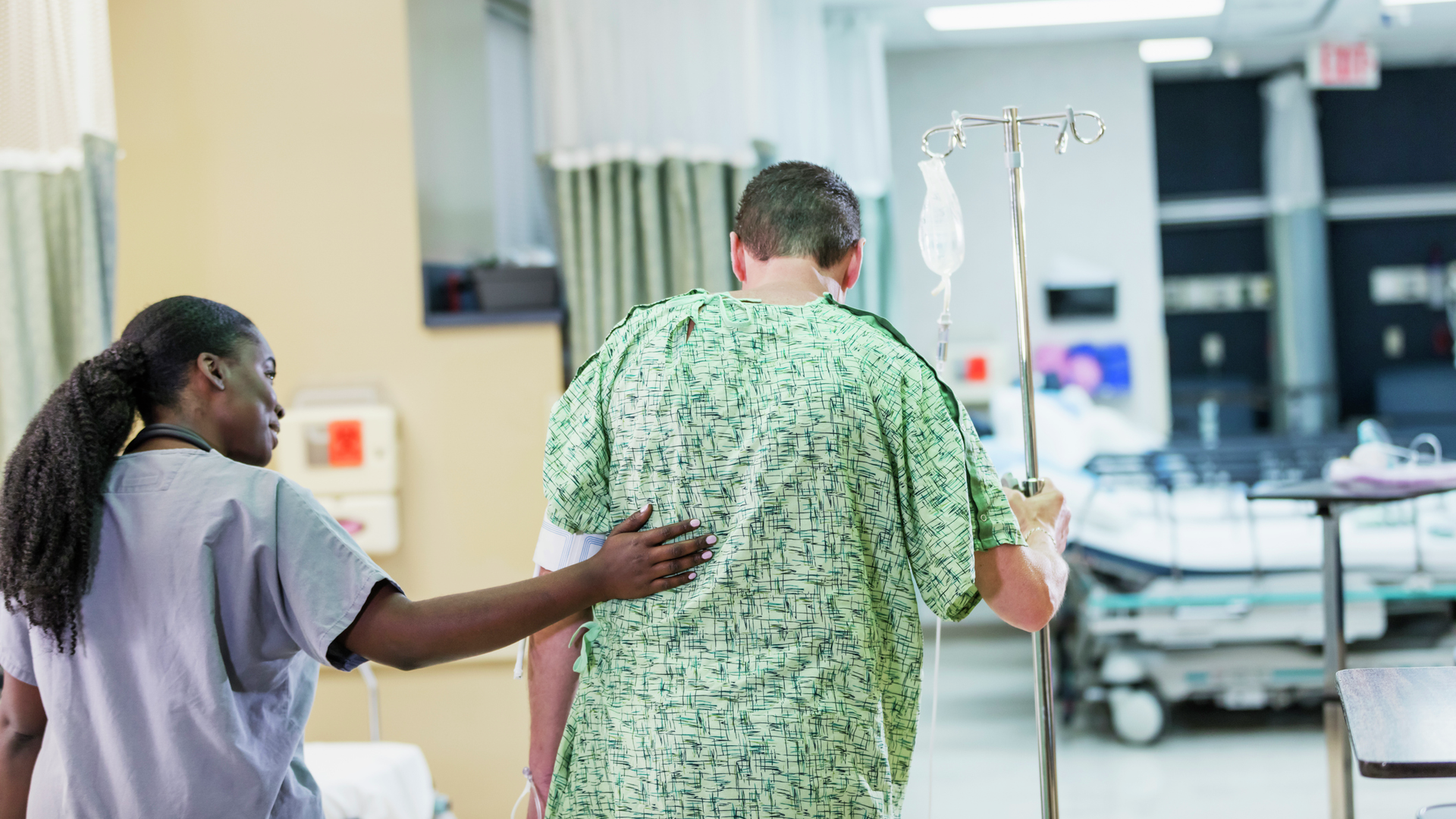 Rear view of a male patient in a hospital gown with IV drip, walking, helped by a nurse in scrubs