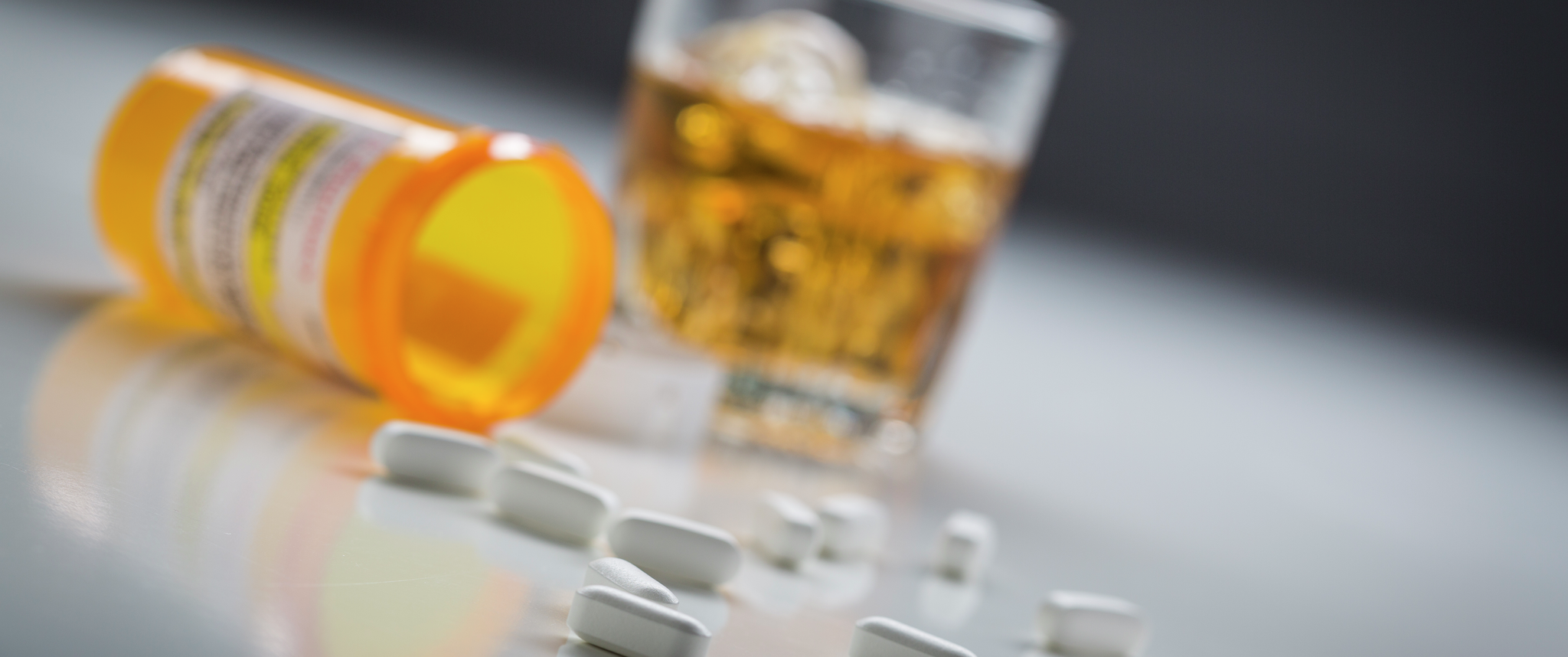 Pills pouring out of a prescription bottle on a surface where a brown alcoholic drink is seen in the background