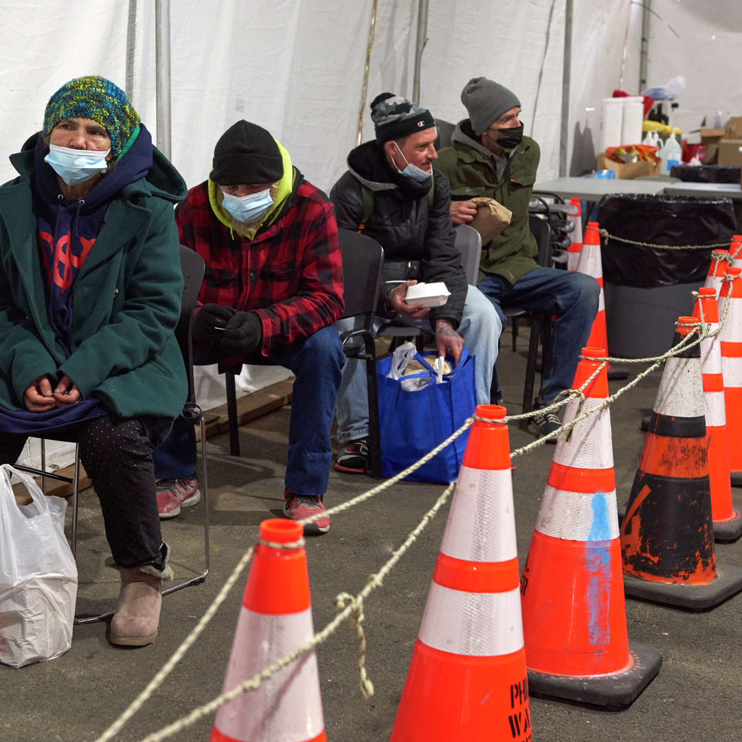 Rose sits in the first of a row of folding chairs all occupied by people wearing winter coats, behind a row of orange traffic cones