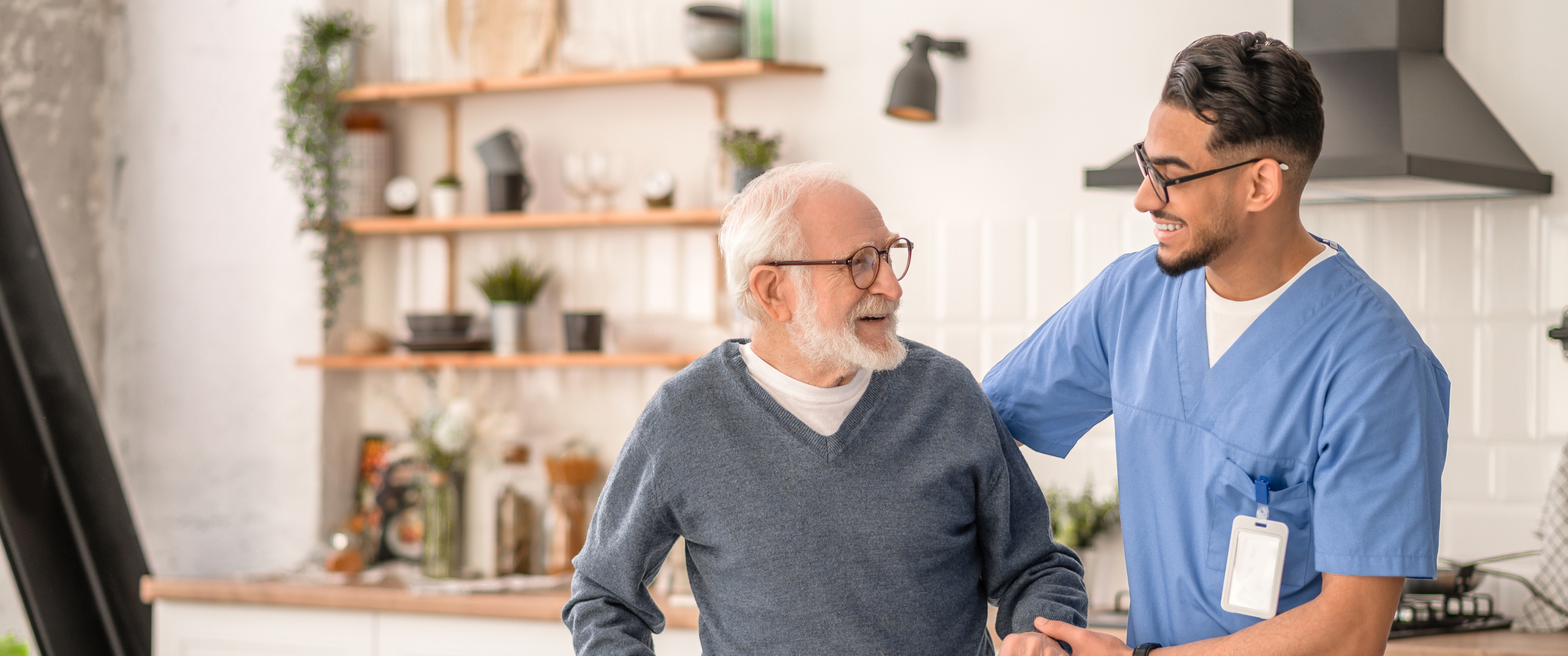 Smiling friendly in-home male nurse in uniform supporting an old man with a walking stick
