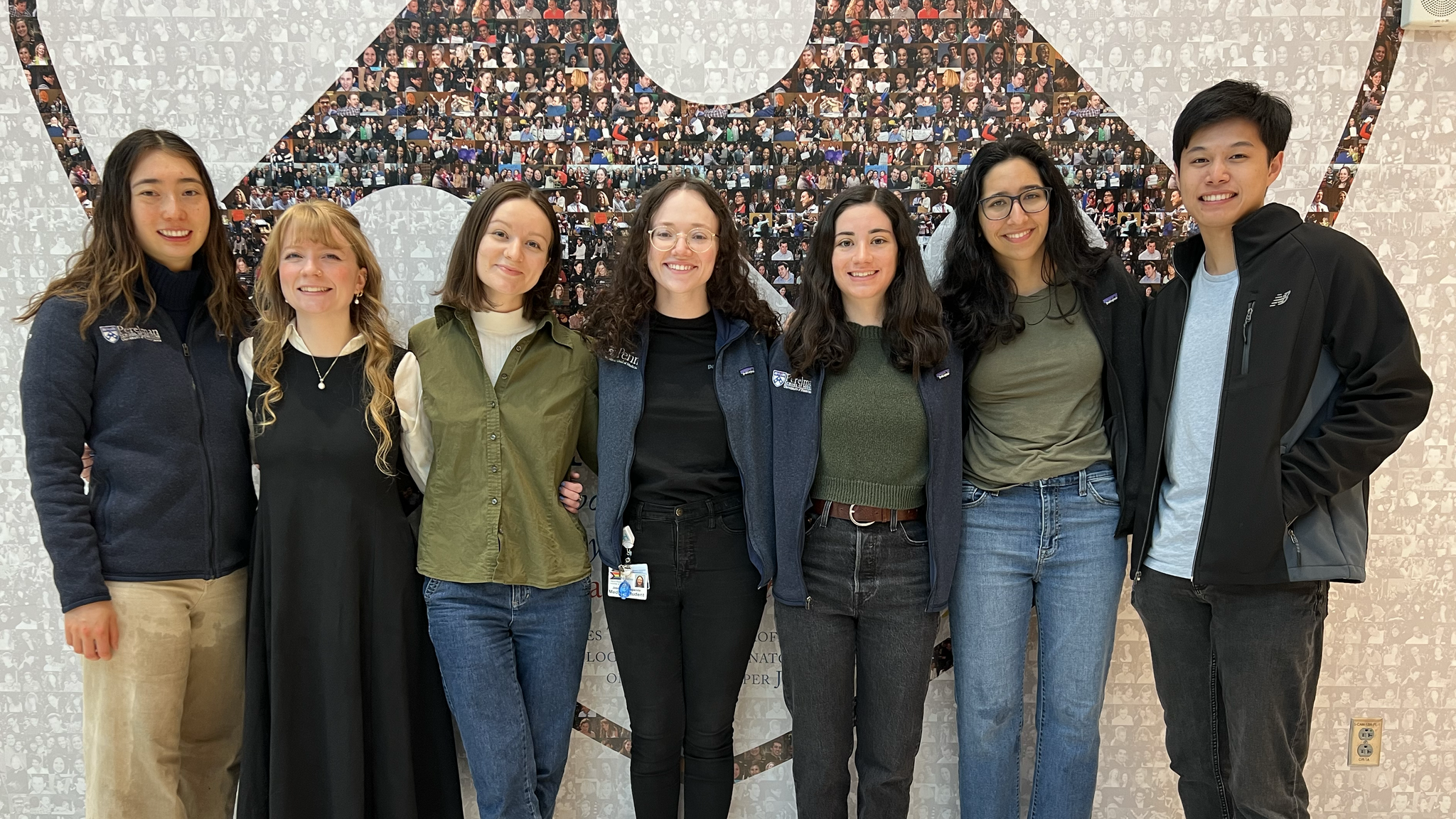 A group photo of medical students Catherine Shen, Elizabeth Whidden, Phoebe Cunningham, Jess Campanile, Sarah Wornow, Isha Thapar, and Jonathan Szeto standing in front of a photo collage in the shape of the Penn shield.