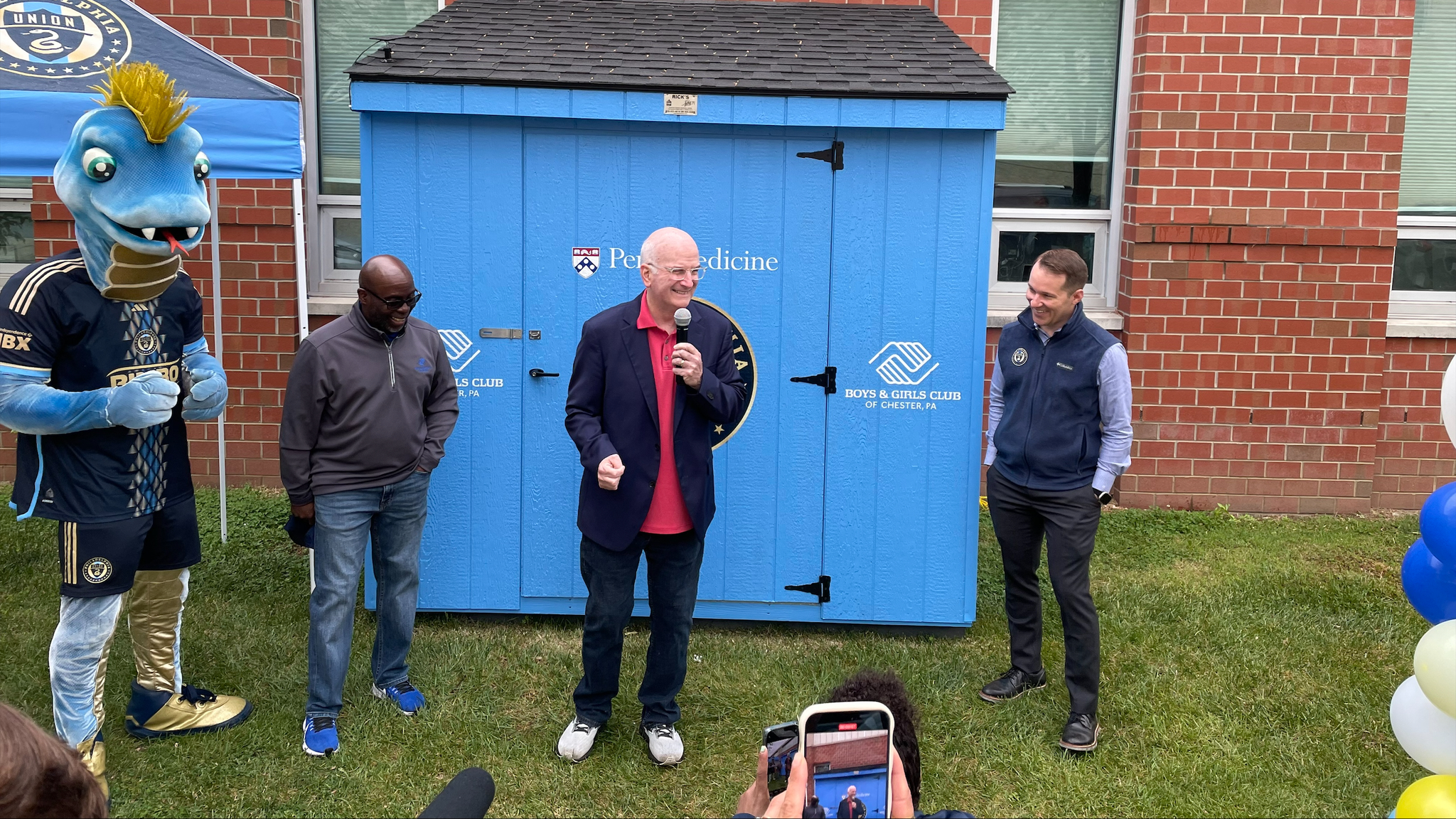 Kevin Mahoney holds a microphone standing in front of a bright blue shed (community food pantry), surrounded by Philadelphia Union officials and mascot Phang