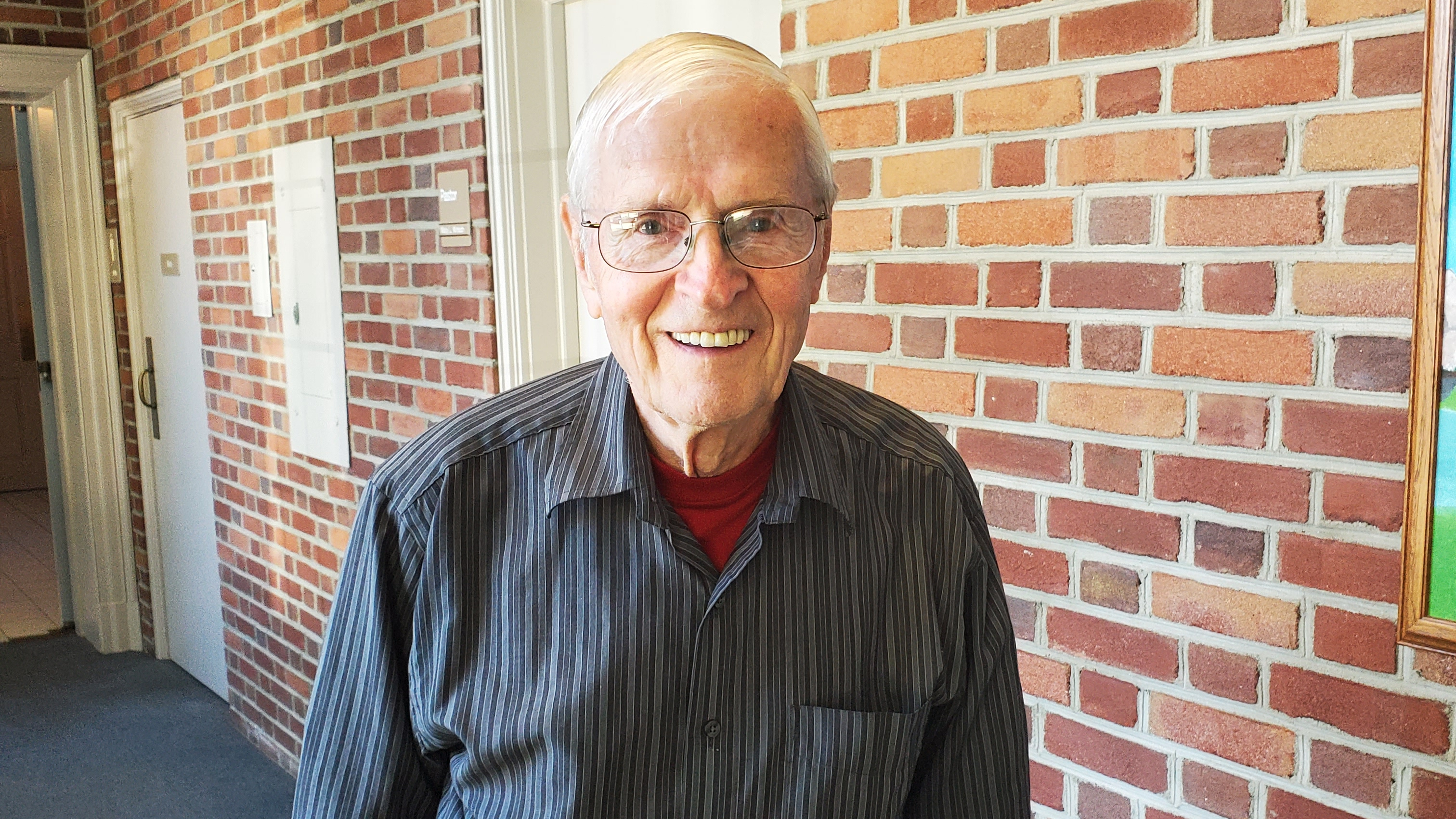Elderly man standing in front of brick wall wearing gray shirt and black pants