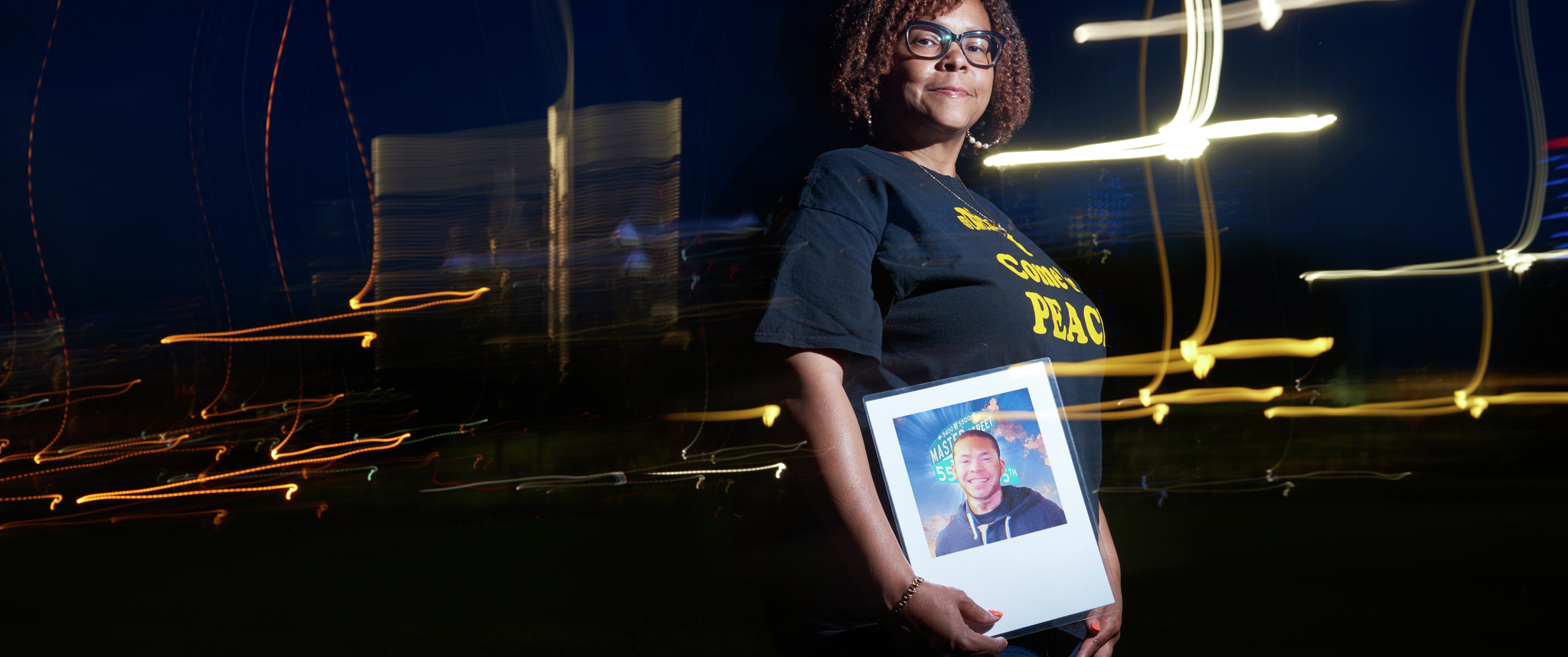 Cheryl Graham-Seay, at night with a blurred city skyline behind her, stands facing the viewer, holding a photo of her son