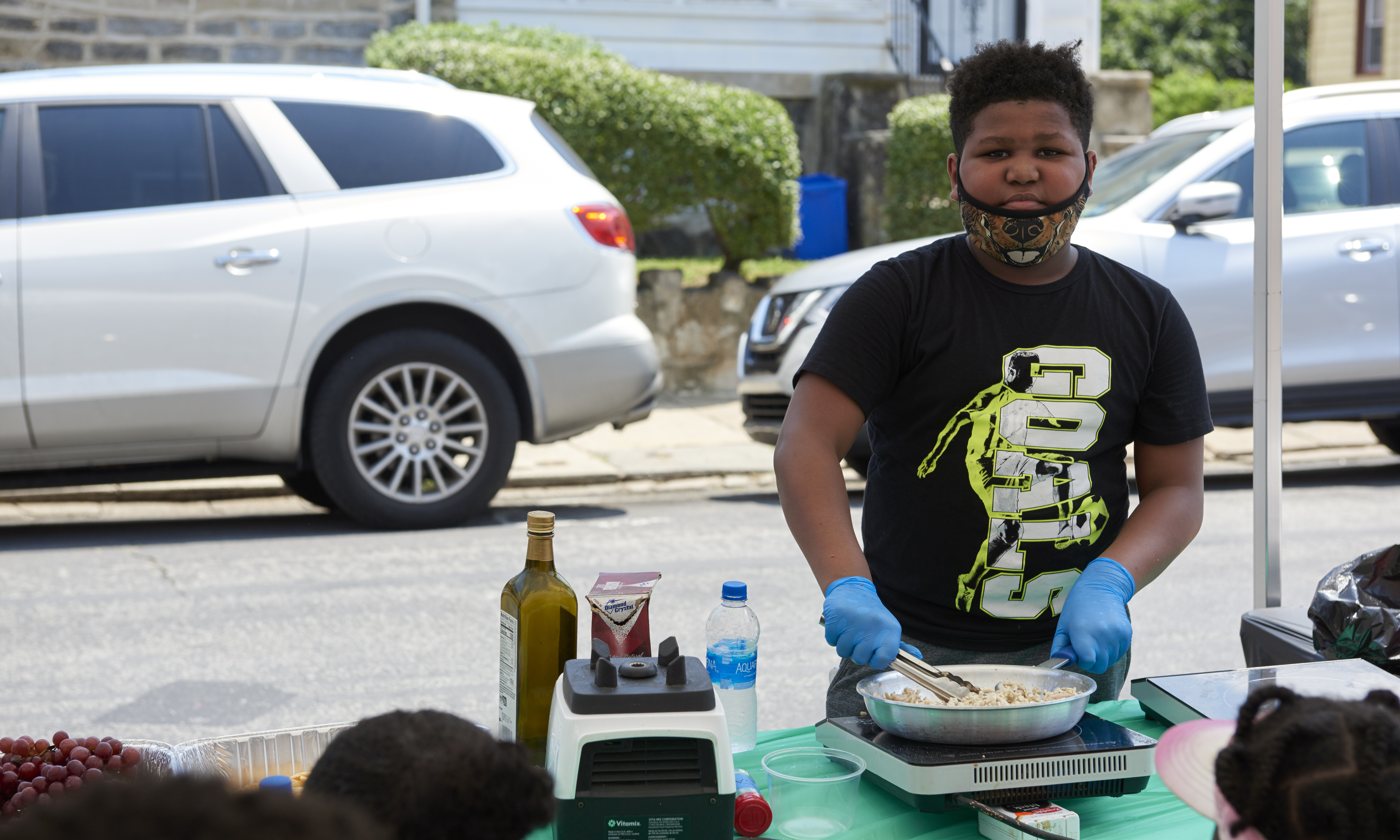 A boy stands at an outdoor table in an urban setting, heating food for a group on a hot plate
