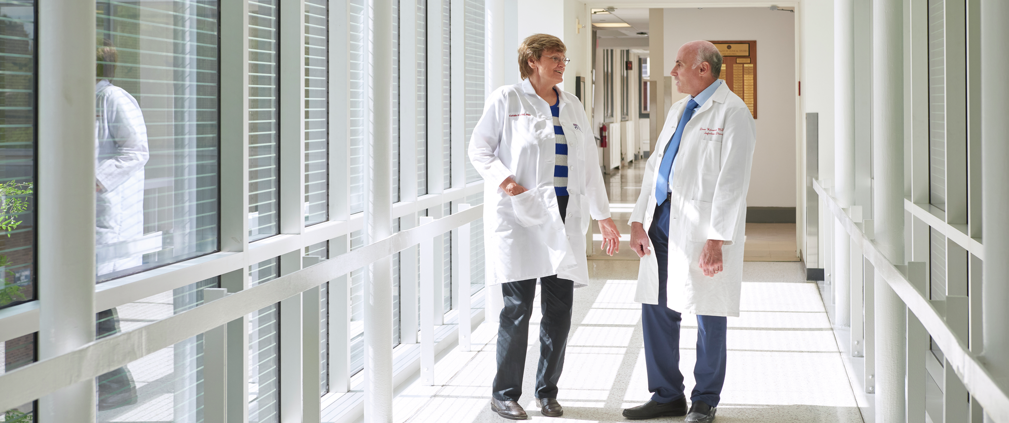 Katalin Karikó and Drew Weissman wearing white coats, talking to one another in a hallway