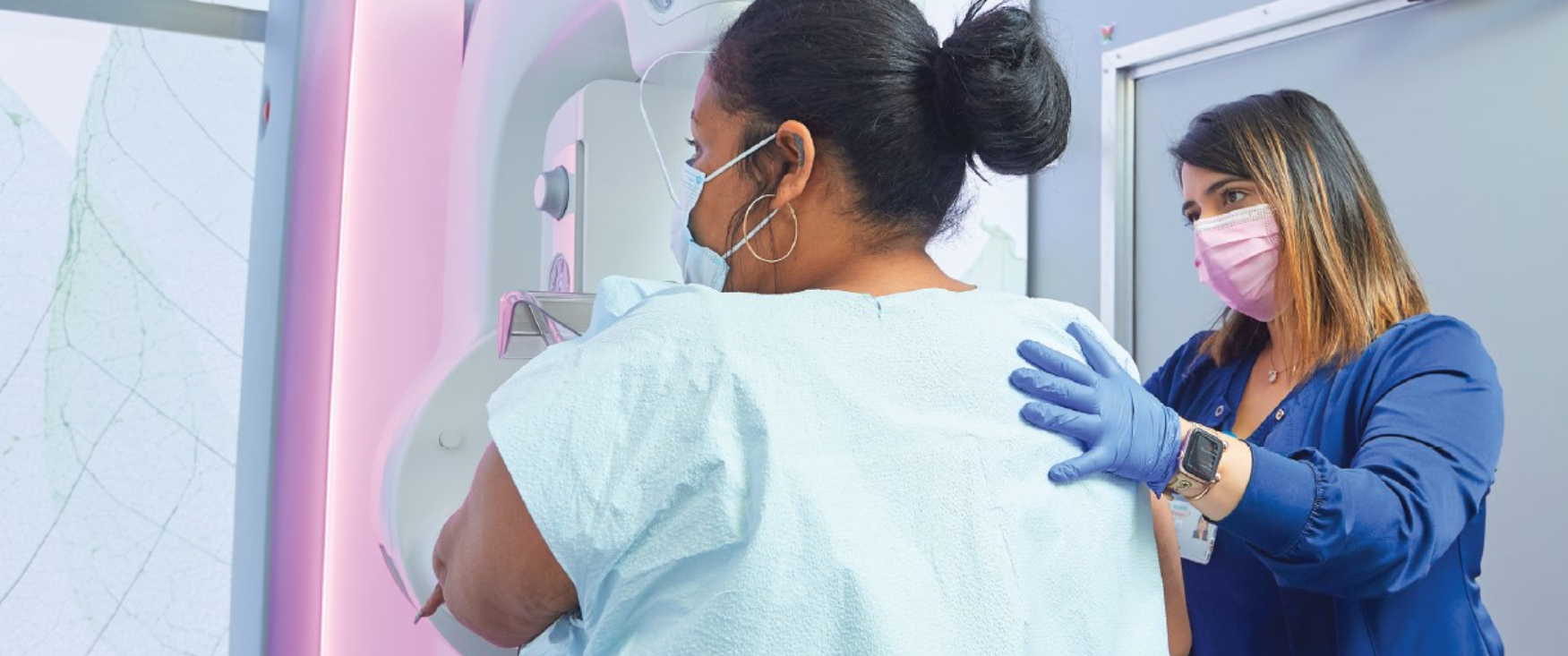 A woman stands with her back to the viewer, her front in a mammography machine, while a technician places a hand on her shoulder