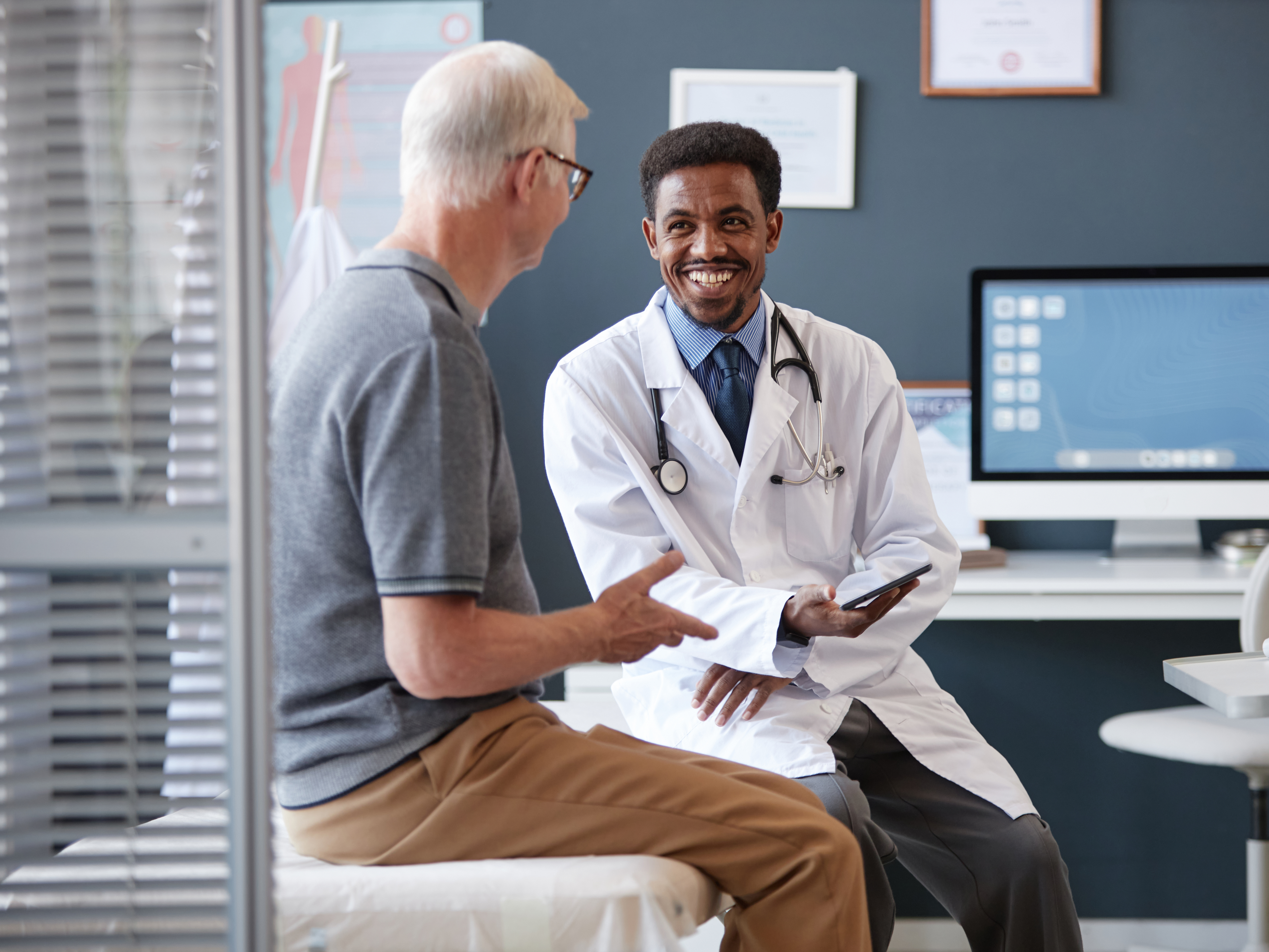 Doctor wearing lab coat and holding tablet while talking to senior patient during consultation.