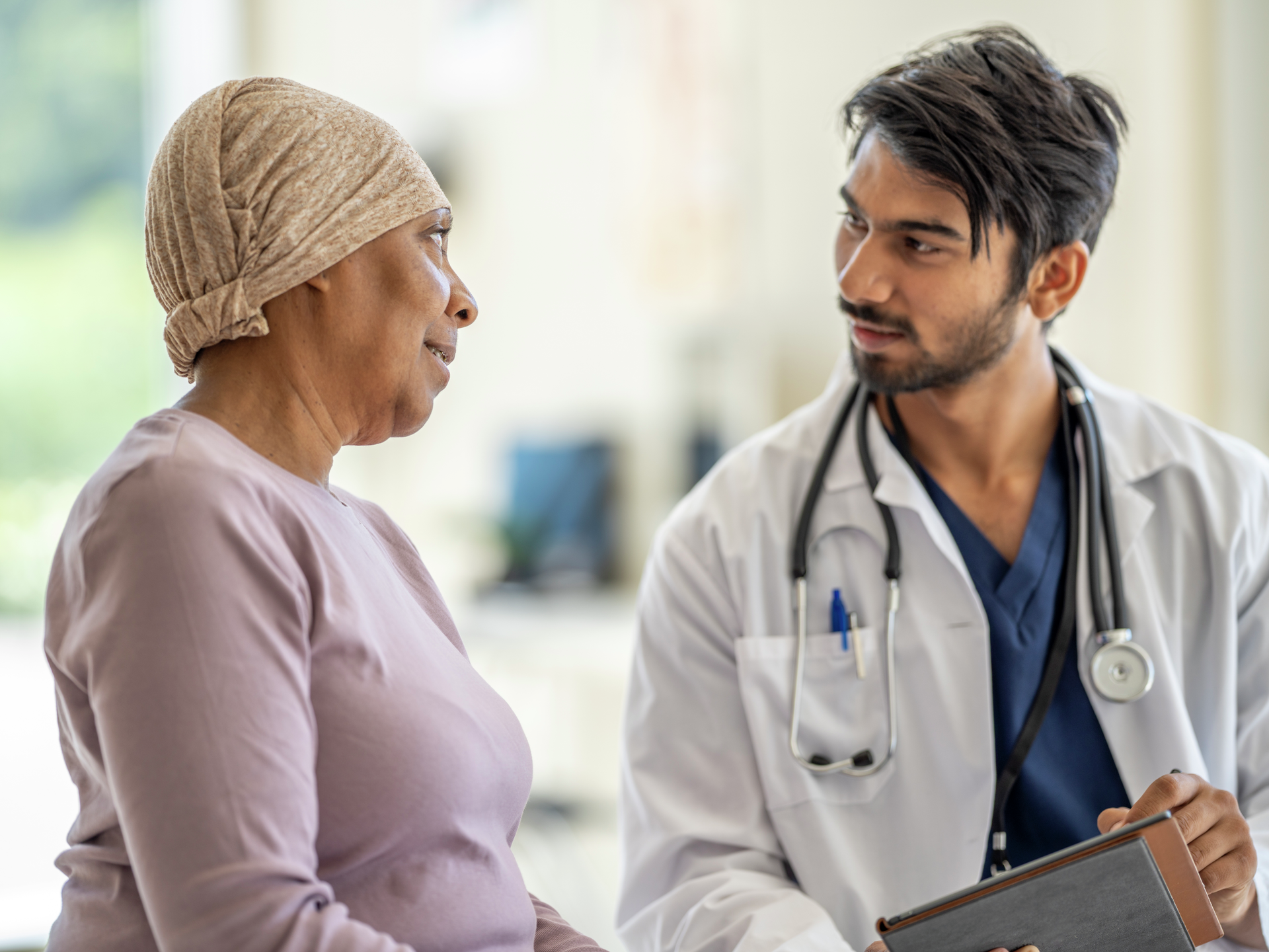 A male Oncologist of Middle Eastern decent sits with a senior patient as they discuss her plan of care. The patient is dressed casually and wearing a head scarf to keep her warm. The doctor is wearing a white lab coat and is holding a tablet as the two meet.