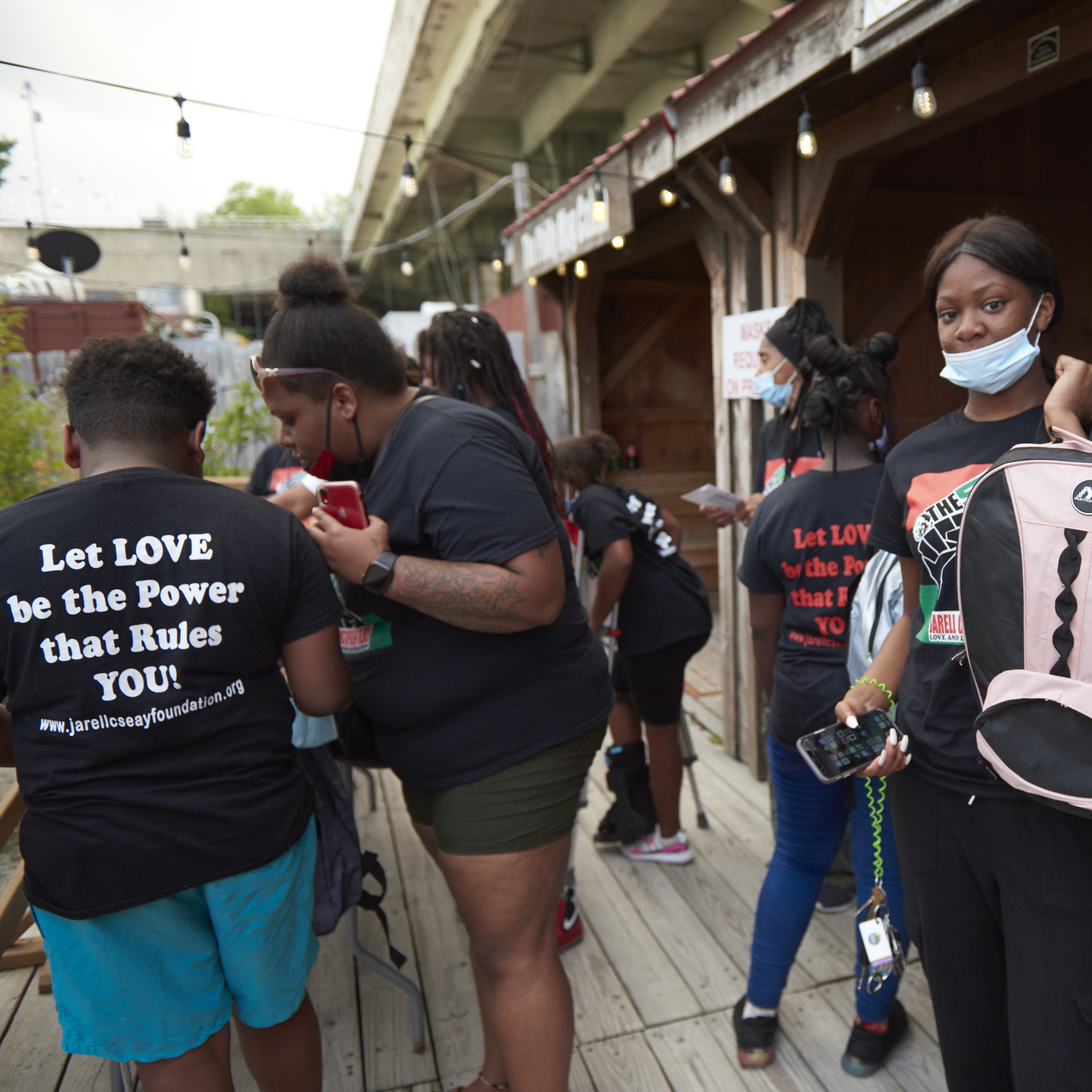 Children and teens gather backpacks, and one child's T-shirt back says "Let Love Be the Power that Rules YOU"