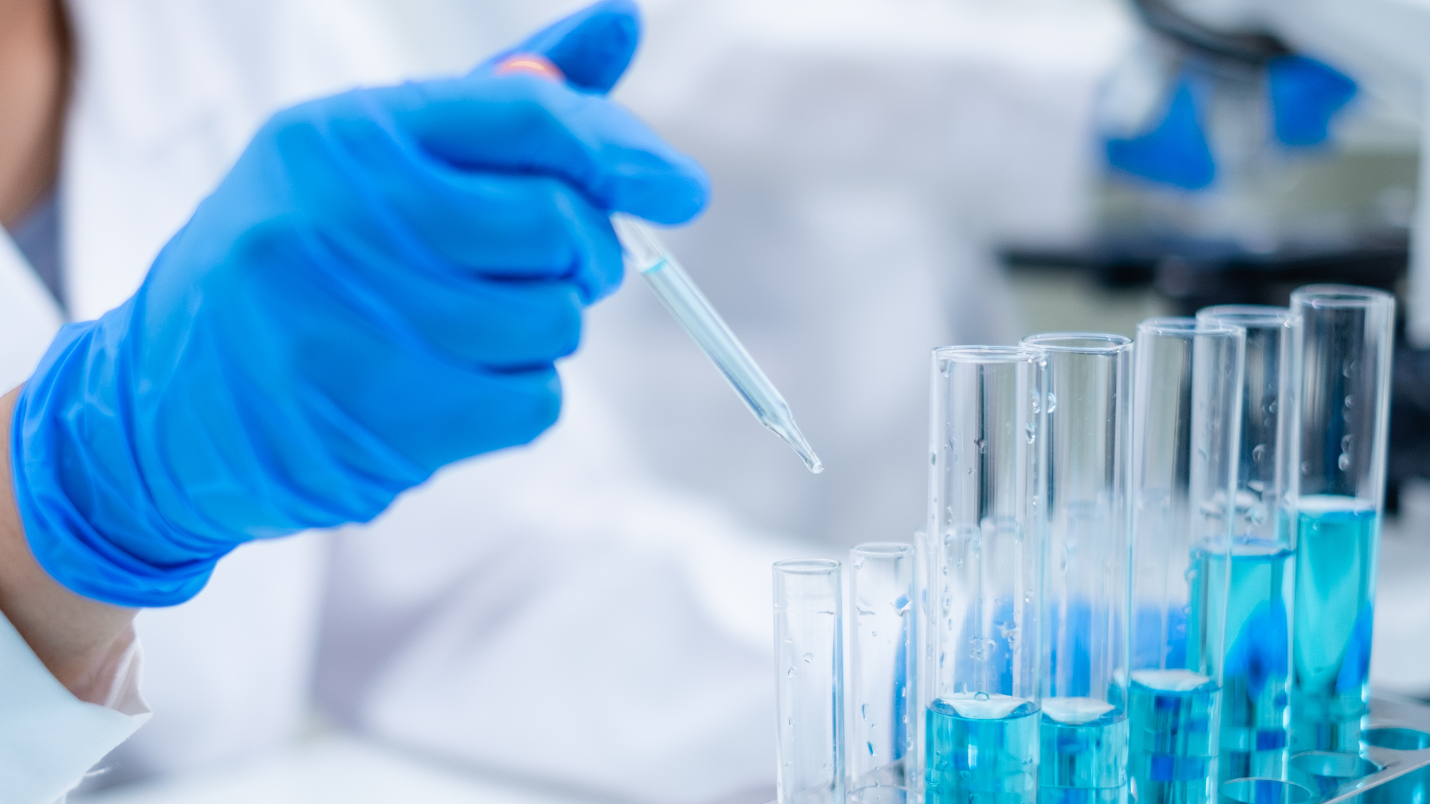 A close up of a scientist’s hands in blue gloves pipetting blue liquid into a test tube