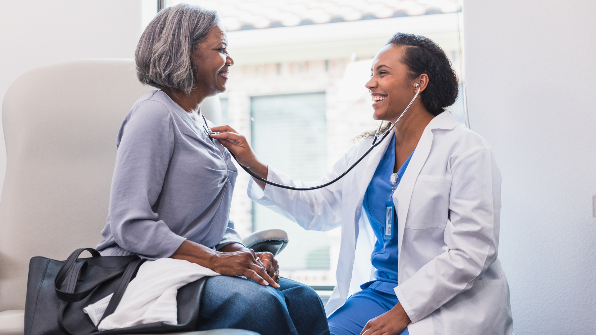 A smiling female doctor listens to a senior female patient's heart during a medical exam