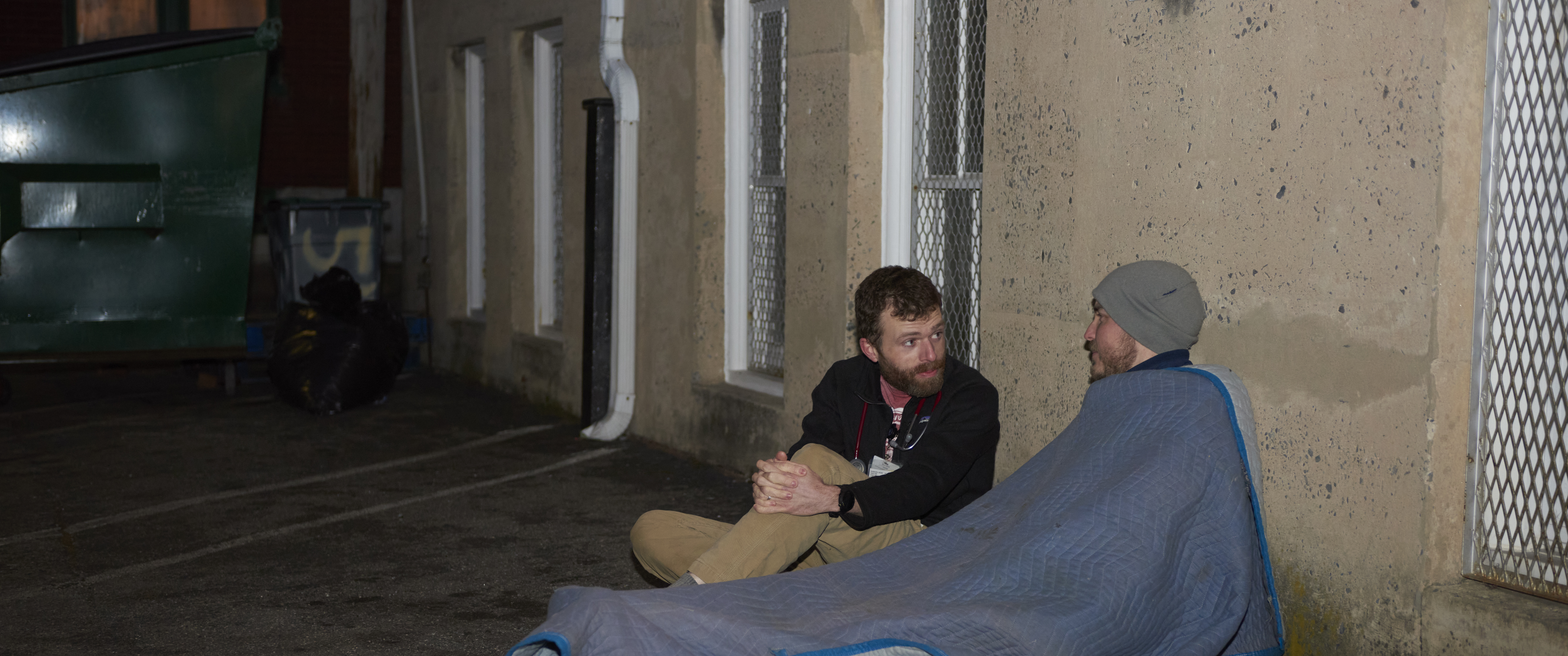 In a parking lot, a doctor sits on the ground speaking with a man lying under a blanket
