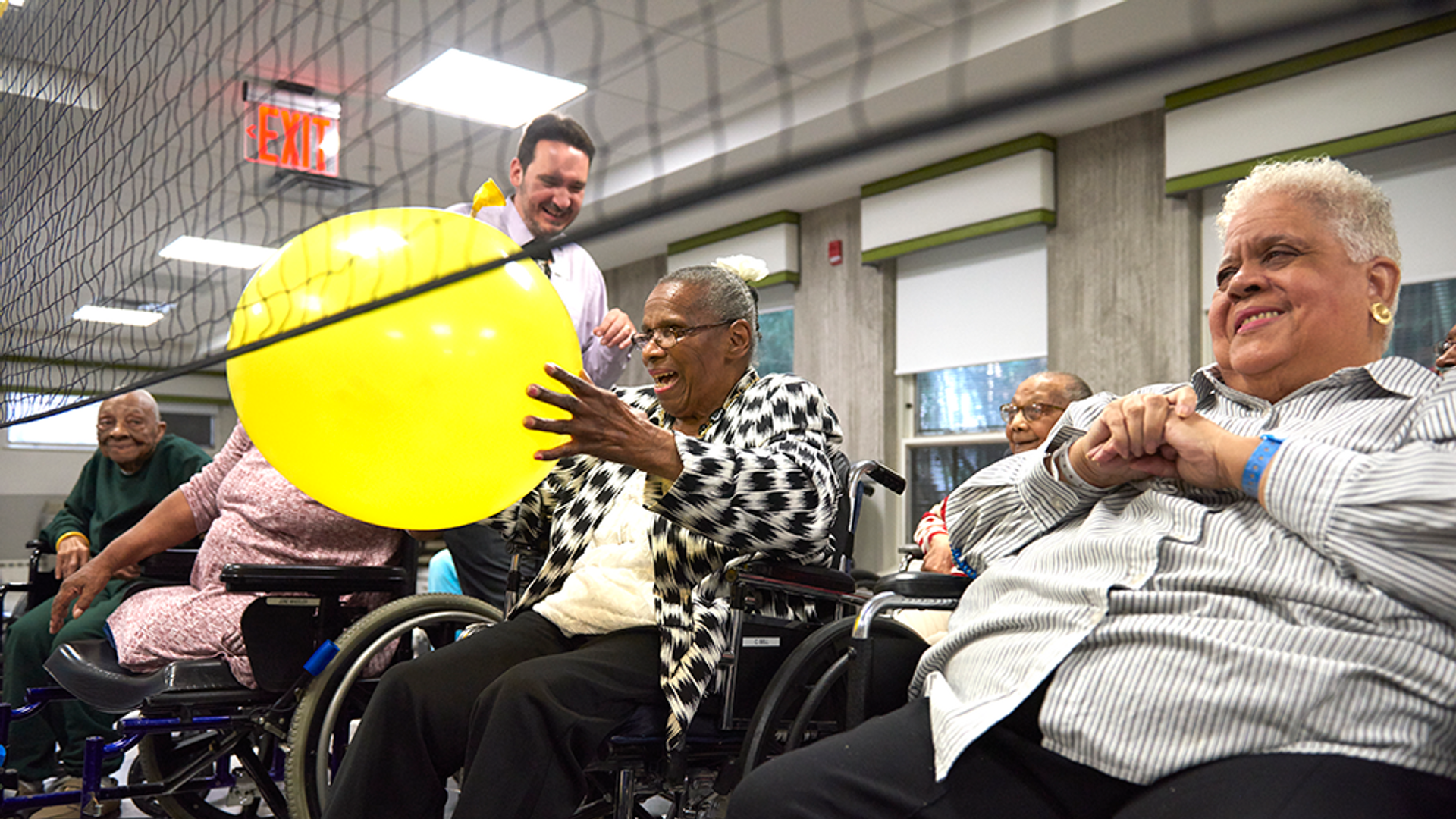A row of senior citizens in wheelchairs sit behind an indoor volleyball net while one holds a large round balloon
