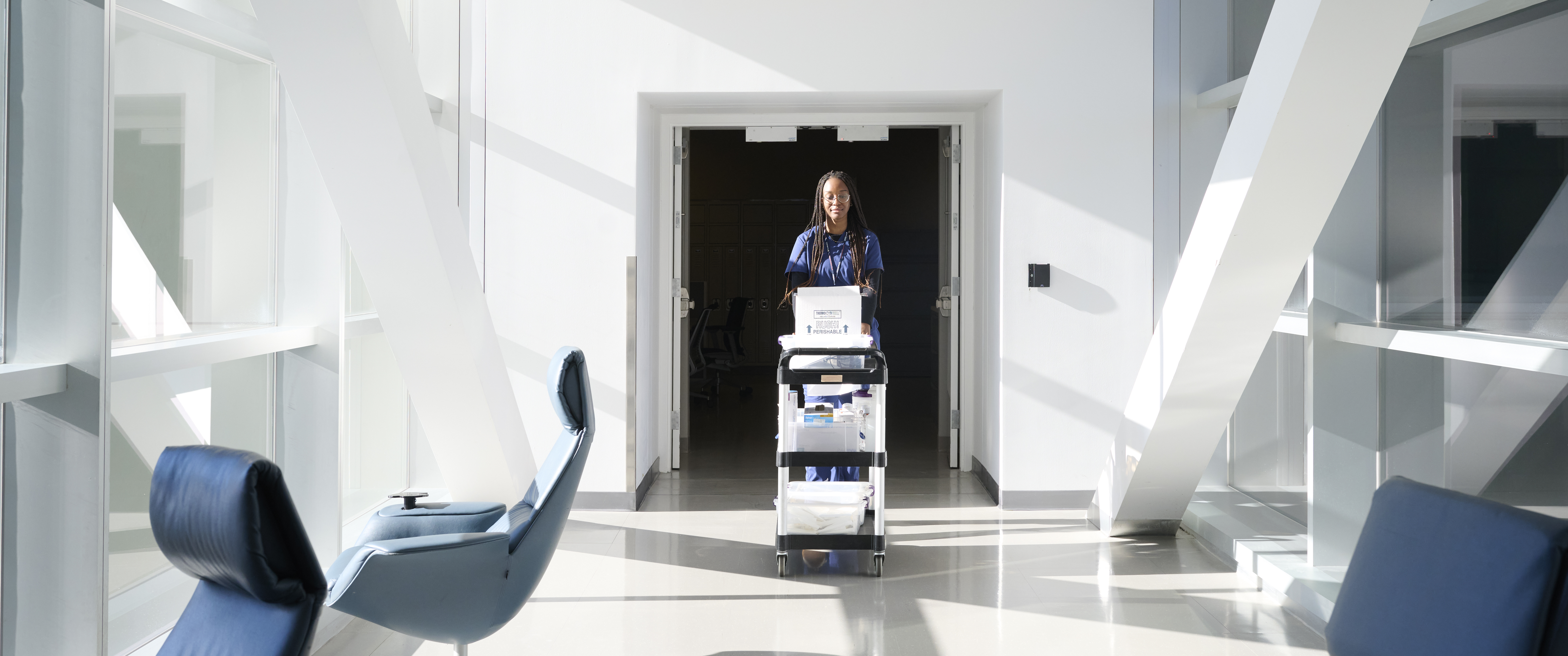 A clinical research staff member pushes a cart through a light-filled hallway, carrying a patient’s CAR T cells and supplies for their infusion