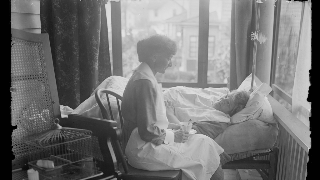 A black and white photo from early in the 20th century depicts a nurse seated at a patient's bedside on a screened-in home porch