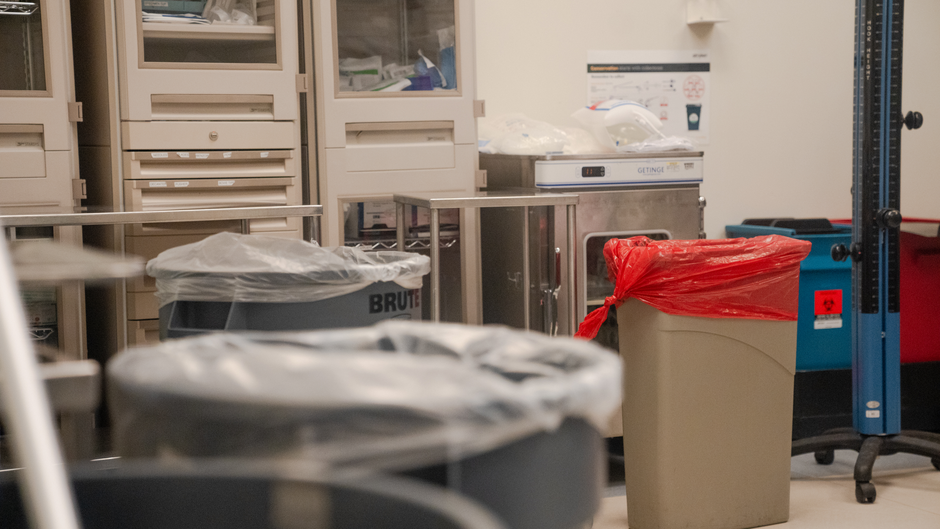 In an empty operating room, a large trash can with a clear plastic bag is seen in the foreground and a smaller can with a red bag in the background.