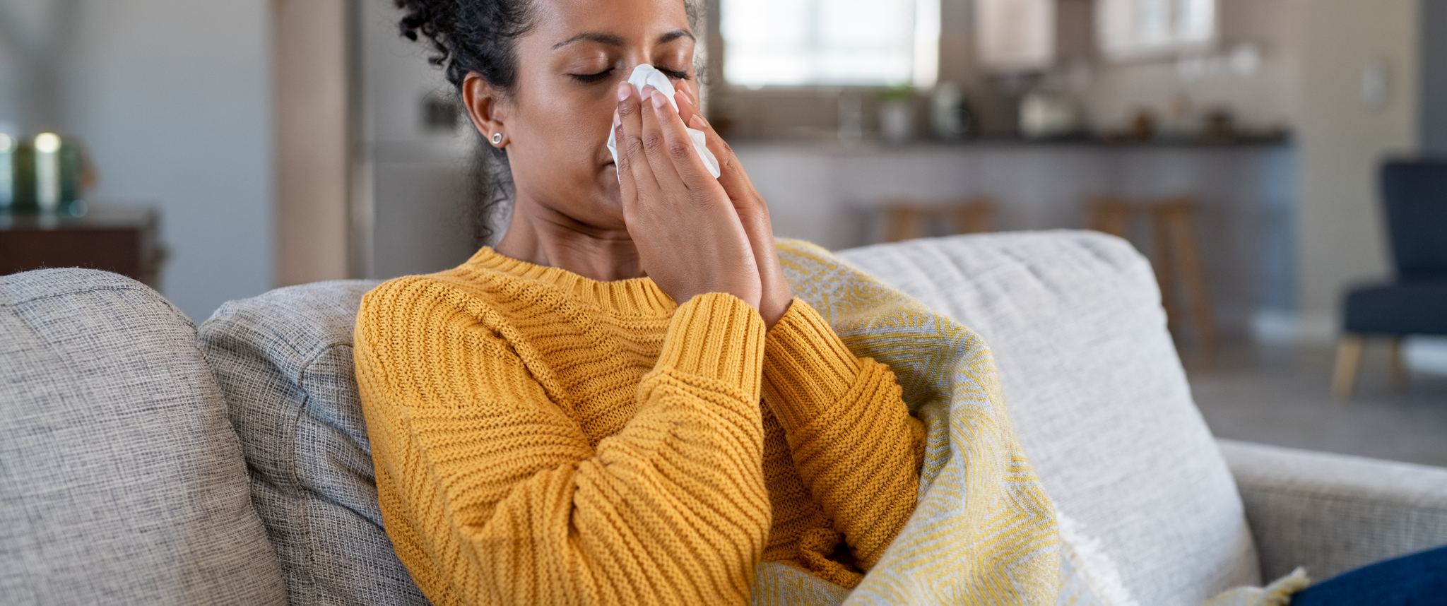 A woman sits on a couch wrapped in a blanket and holds a tissue up to her nose