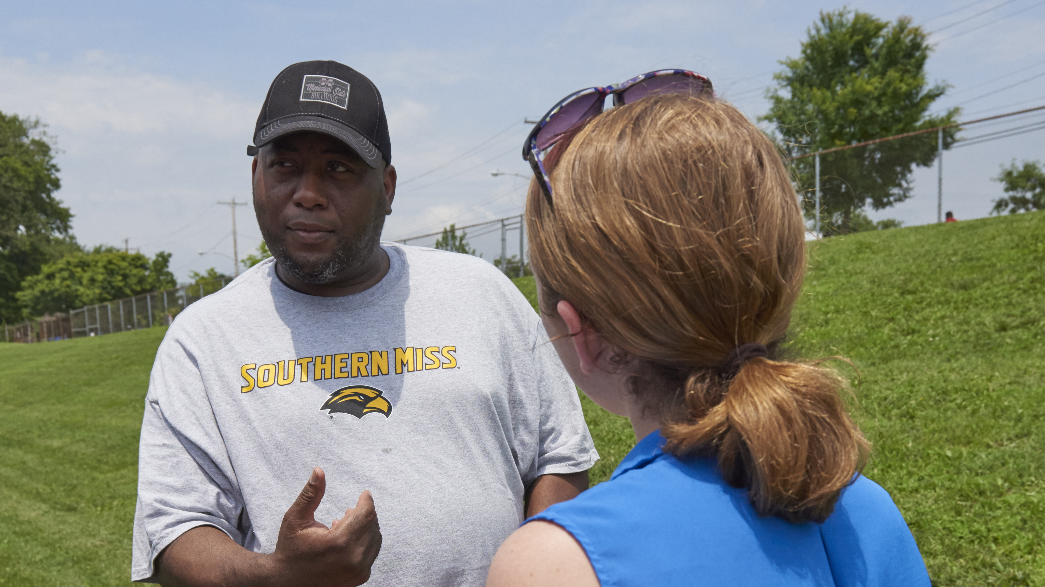 A Black man looks to the side as if contemplating a question while in conversation with a young woman who holds a FIT test kit envelope
