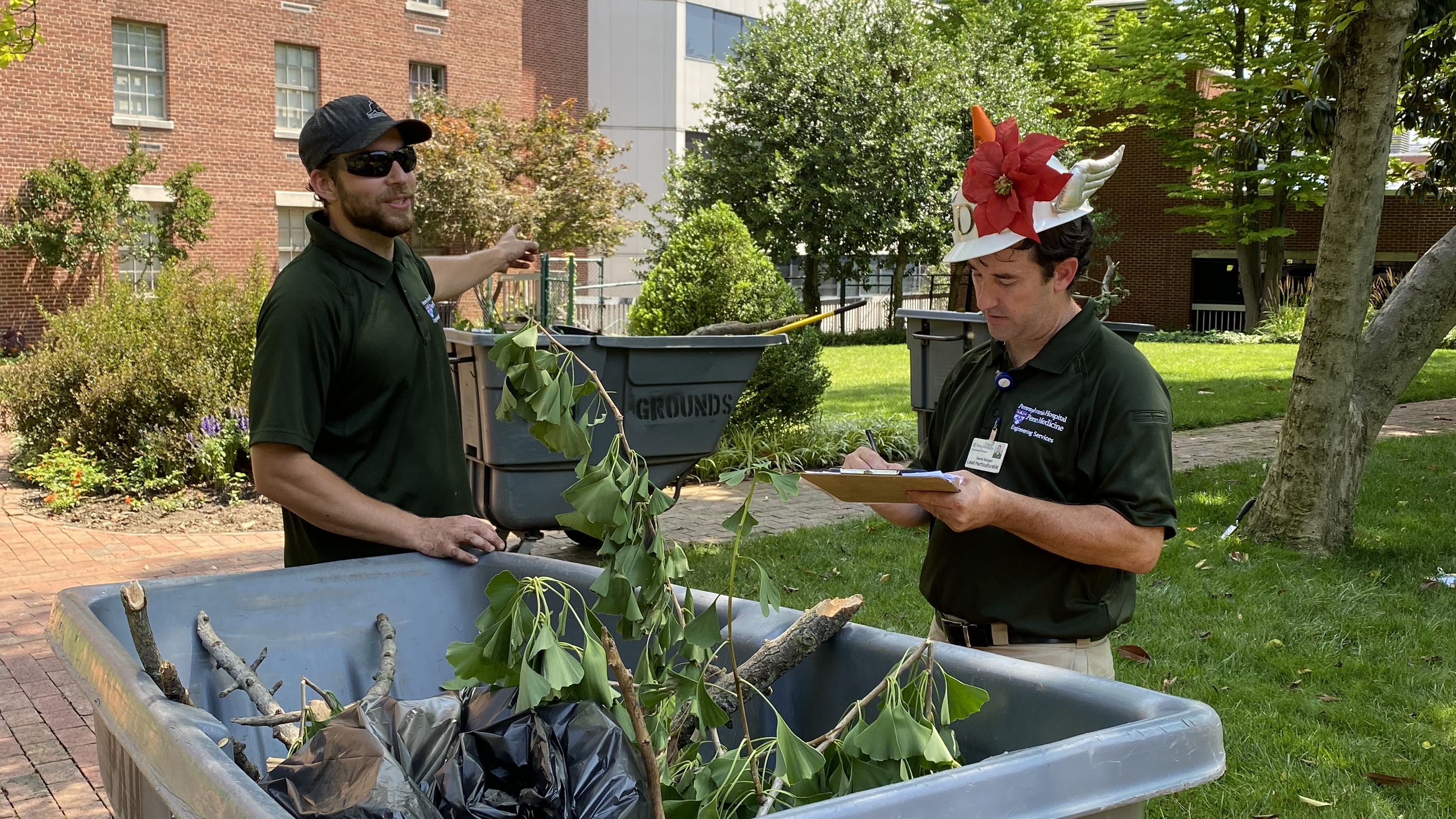 Blake Sieminski stands in front of a cart filled with weeds and tree branches in a Pennsylvania Hospital courtyard, standing next to Dan Bangert who is looking at a clipboard