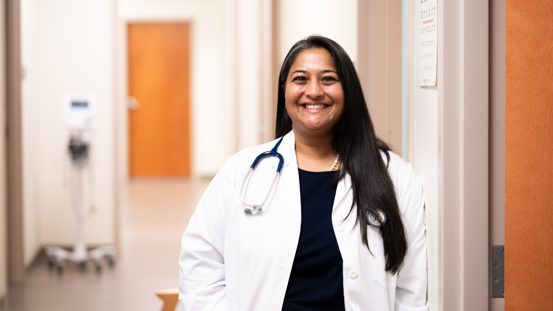 Aileen John, smiling in her white coat and stethoscope, leans against the wall at the Penn Family Medicine West Chester practice