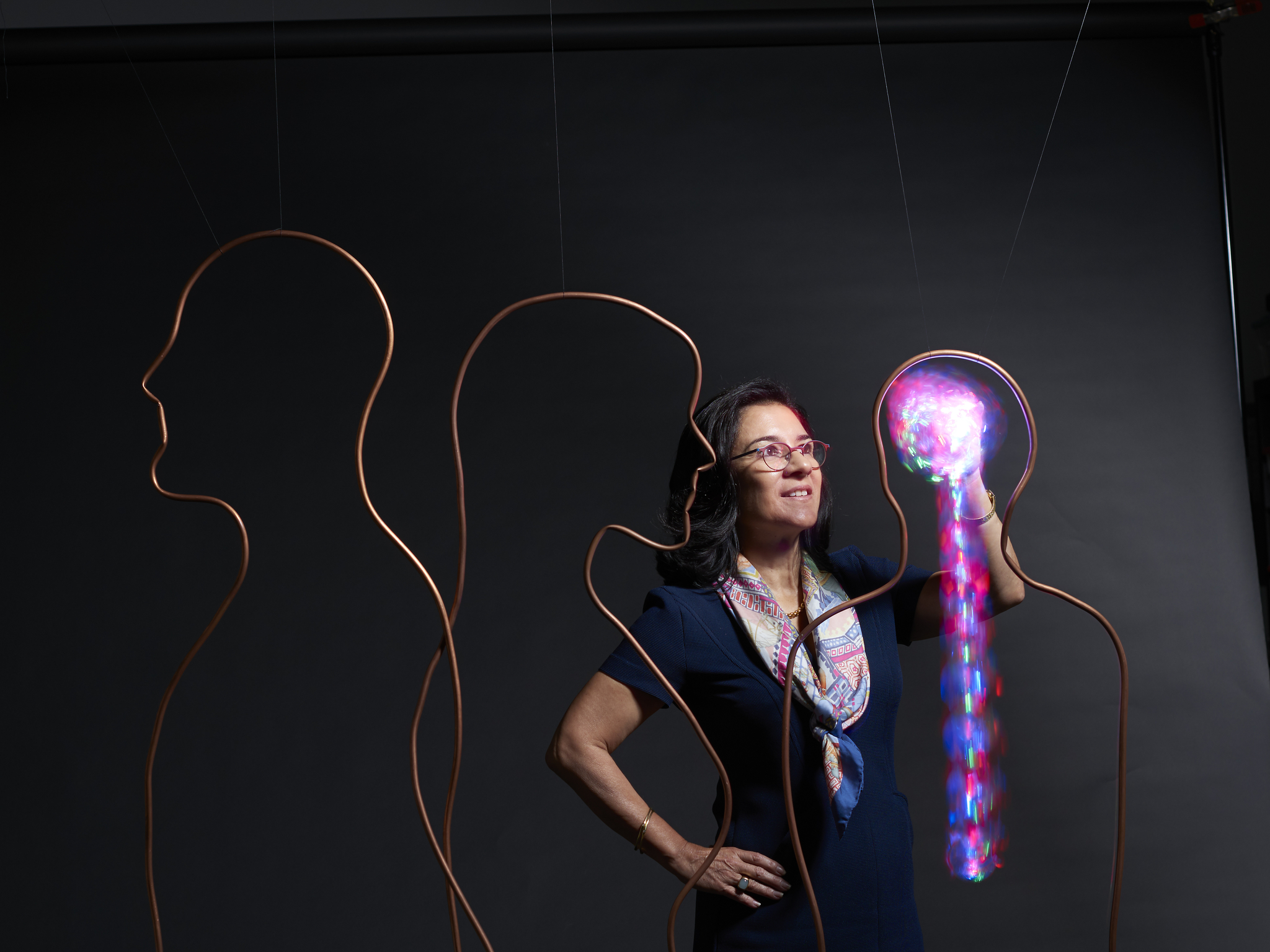 Maria Oquendo stands with one hand appearing enmeshed in a brain shape made of light, within and surrounded by abstract outlines of human silhouettes