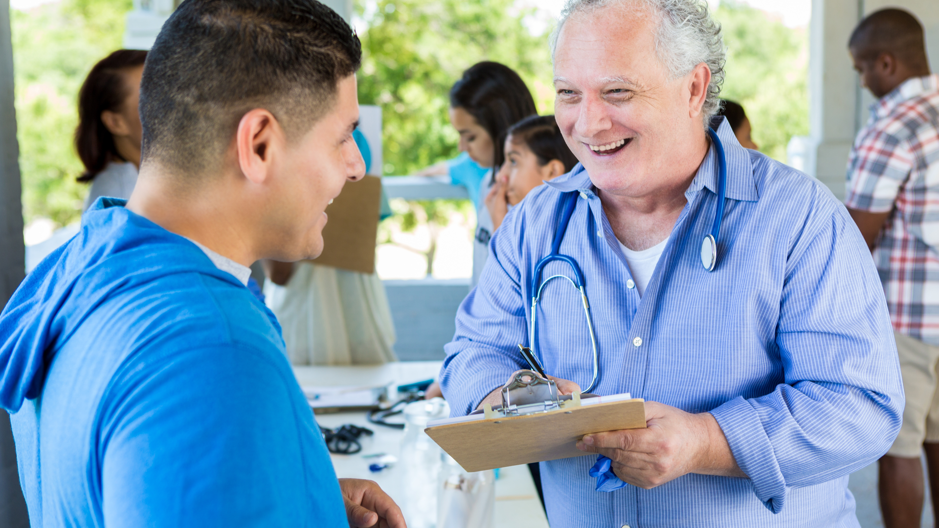Senior Caucasian male doctor talks with mid adult Hispanic male patient at outdoor health fair. The doctor is collecting information from the patient.