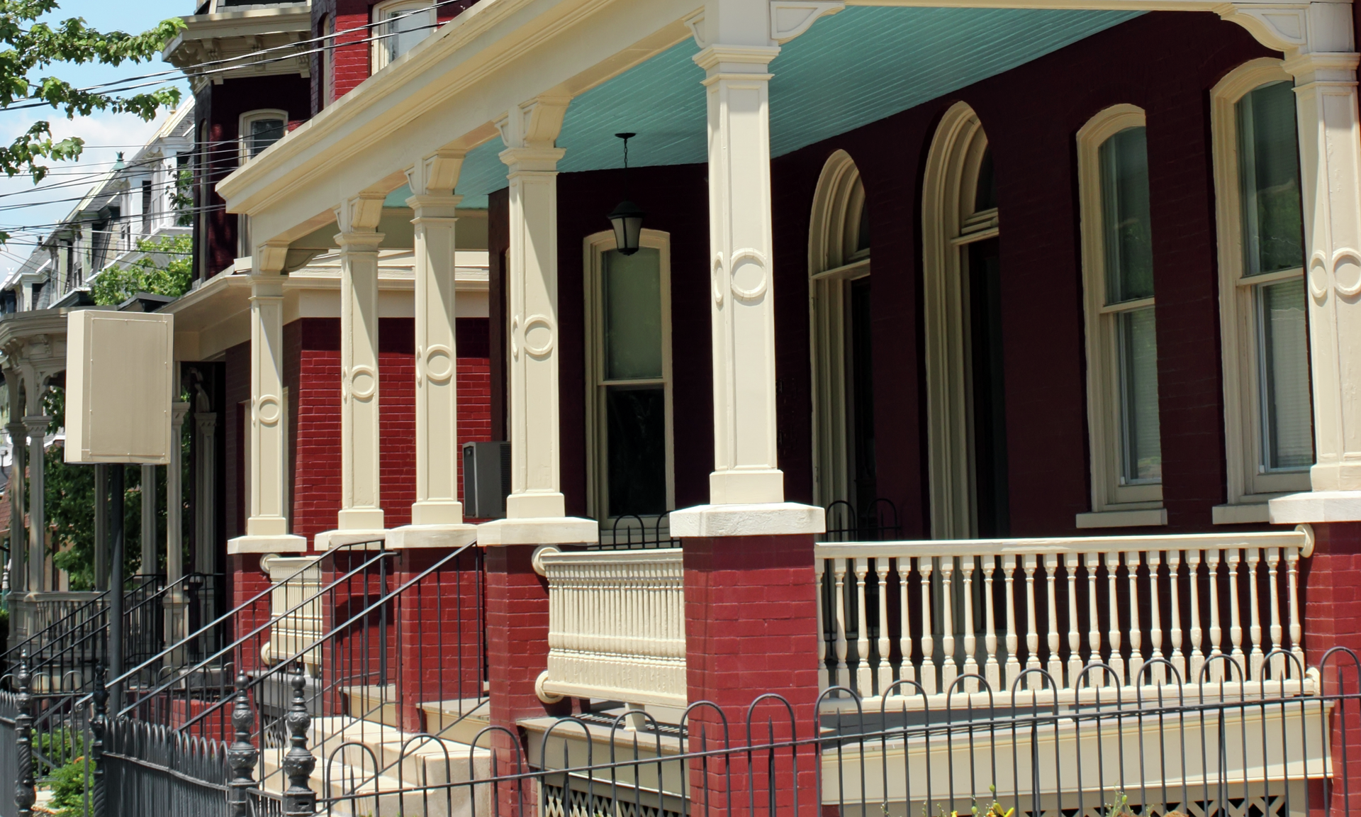 Row of red brick Victorian-style townhouses with bay windows and covered porches, framed by green trees under a blue sky.