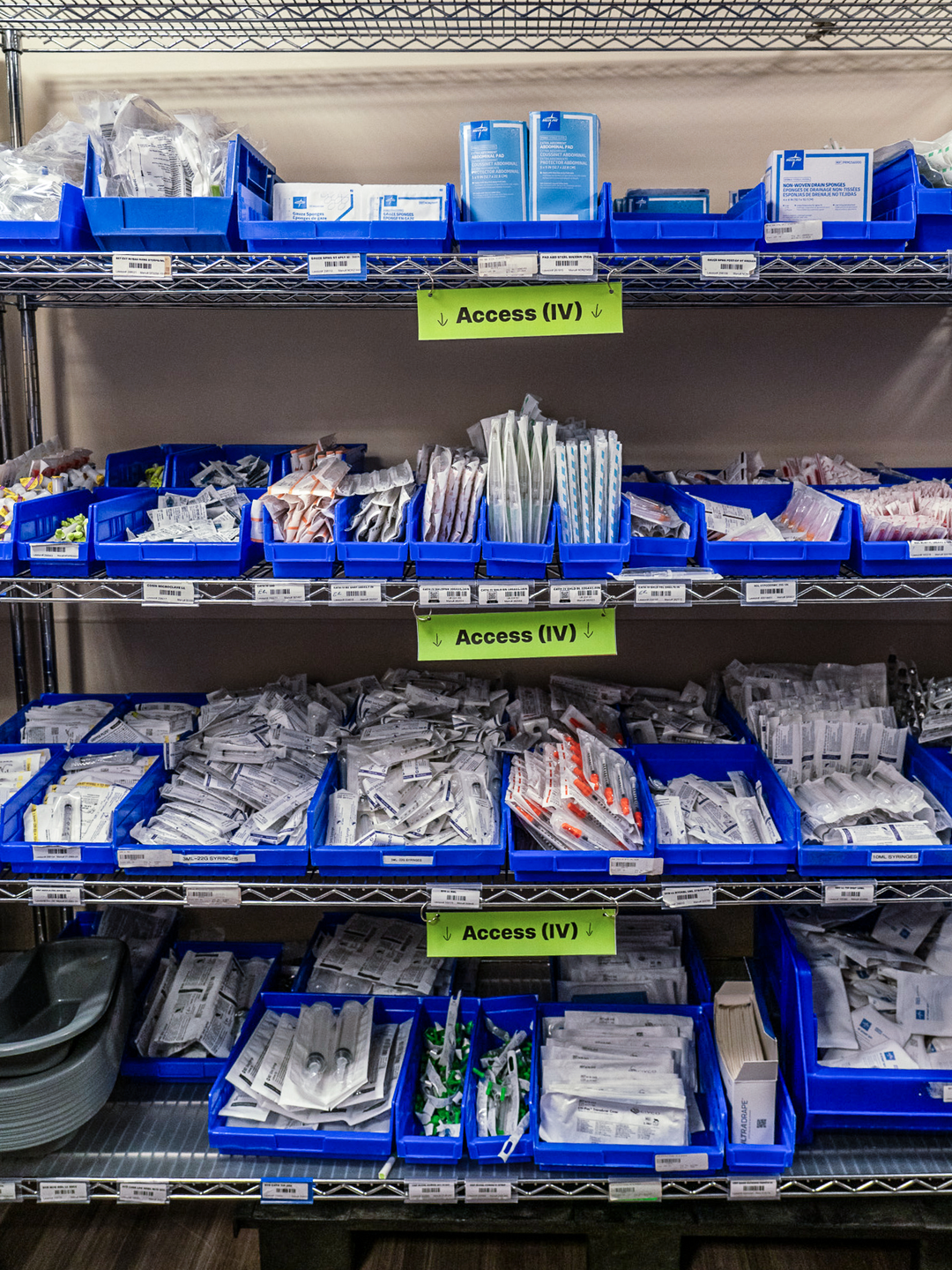Close-up image of a metal shelf with open blue bins filled with medical supplies. A label on each shelf says Access (IV) 