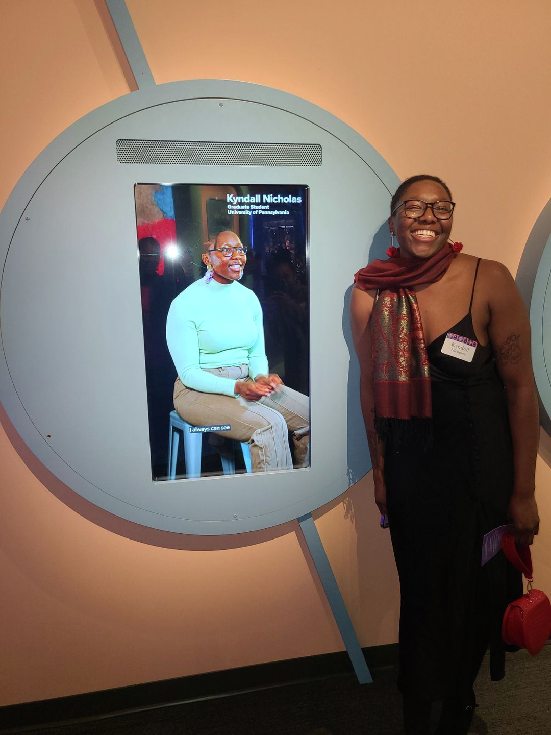 Kyndall Nicholas smiles big standing next to a large video display of herself at the Franklin Institute