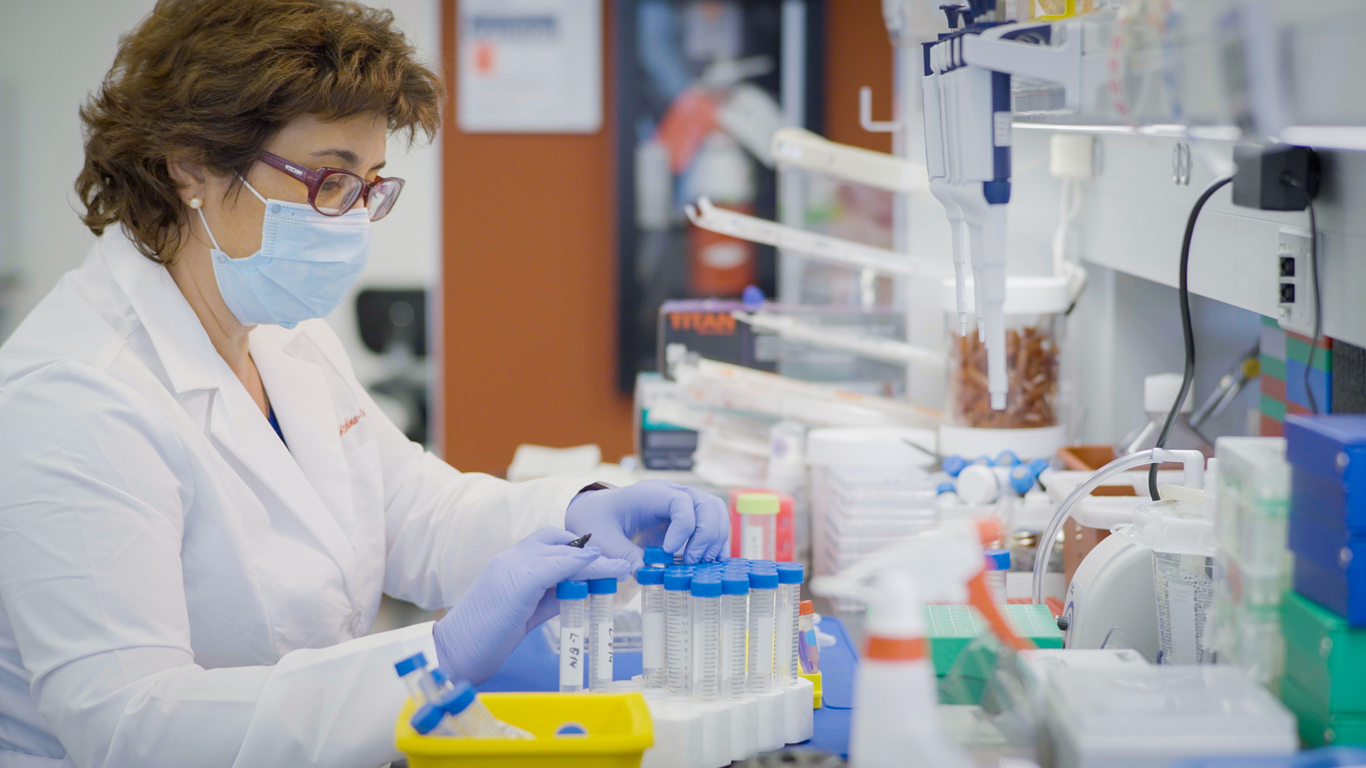 A woman wearing a lab coat, mask, and gloves, works at a lab bench doing mRNA research