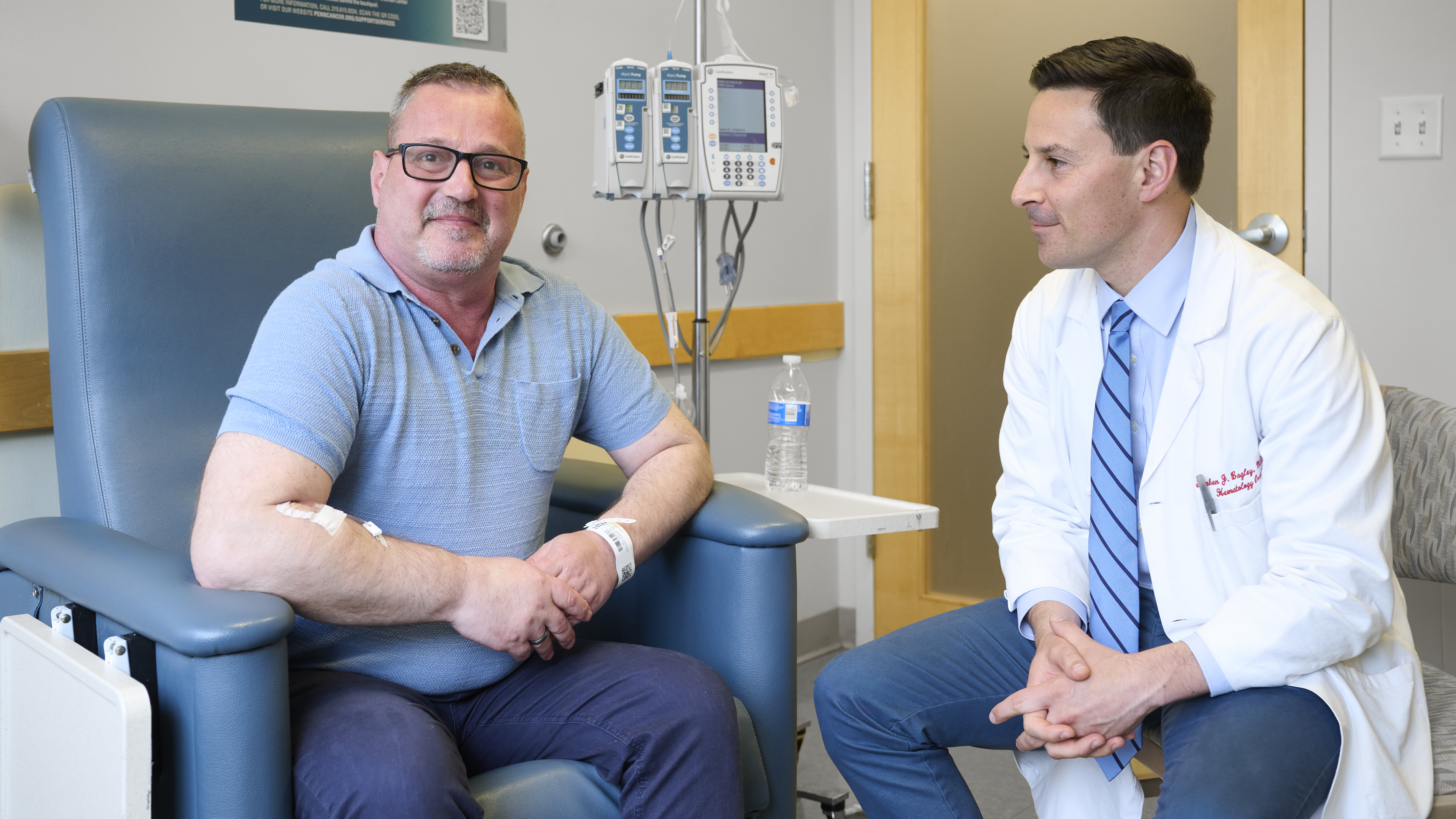 Eduart Cuka, sitting up proud and happy in an infusion chair, with Stephen Bagley, MD, at his side, looking at him 