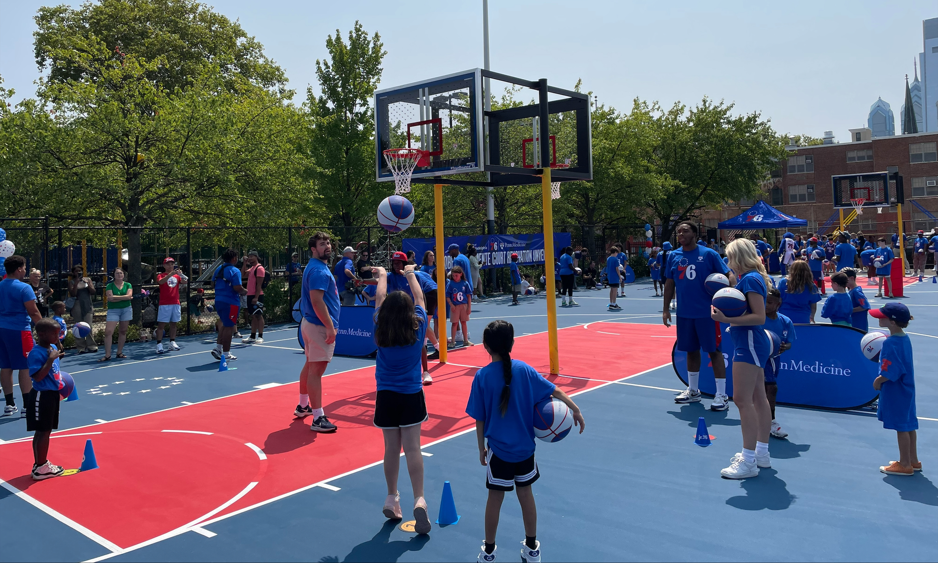 A girl shoots a basketball with families gathered, in Philadelphia Sixers t-shirts, on the new basketball court in Roberto Clemente Park