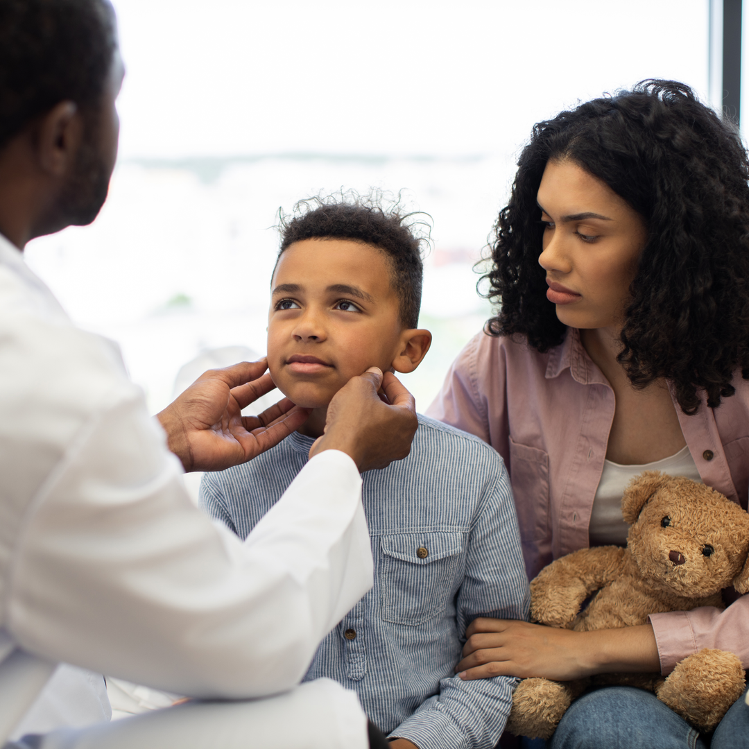 African American male doctor examines young sick African boy's neck lymph nodes. Mother sits beside comforting him with stuffed toy.