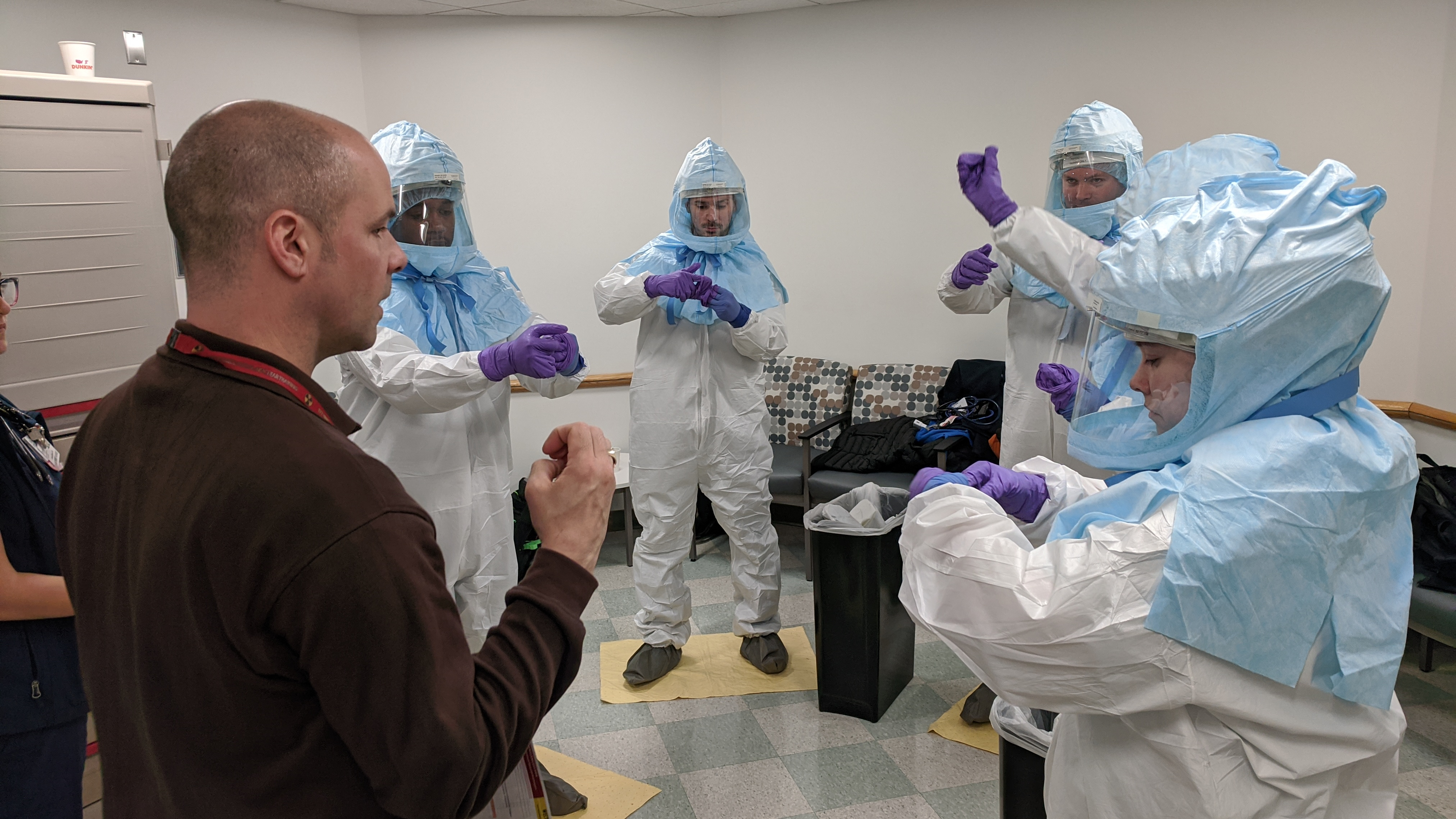 A group of people in personal protective equipment remove gloves, instructed by another person