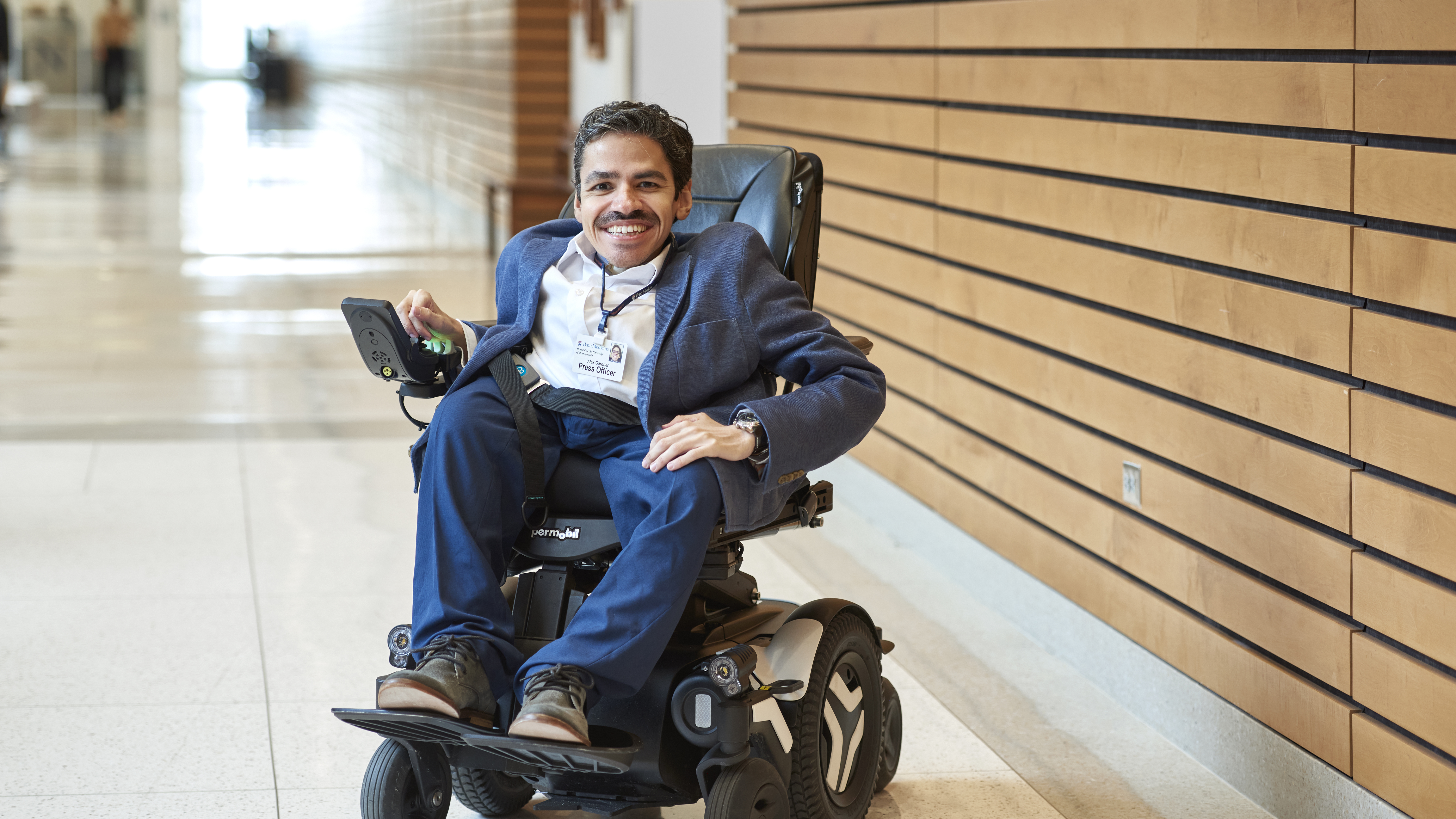 Alex Gardner, seen in a full body photo sitting in a wheelchair, relaxed and smiling, with his right hand on the chair's control panel