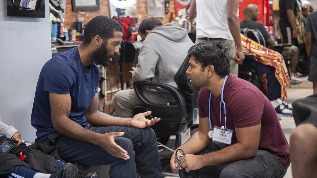  A bearded Black man and a male medical student converse in a barbershop